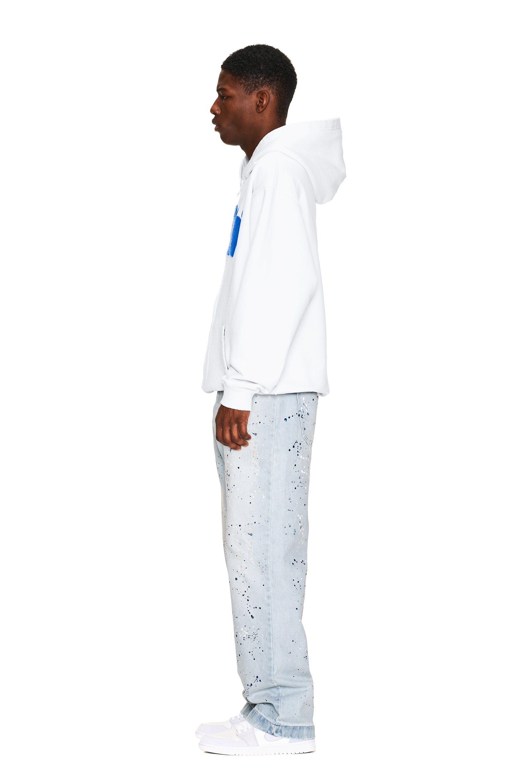 A young man stands in profile against a white background, wearing the 6PM LOGO ZIP HOODIE WHITE, light blue splatter-painted jeans, and white sneakers.