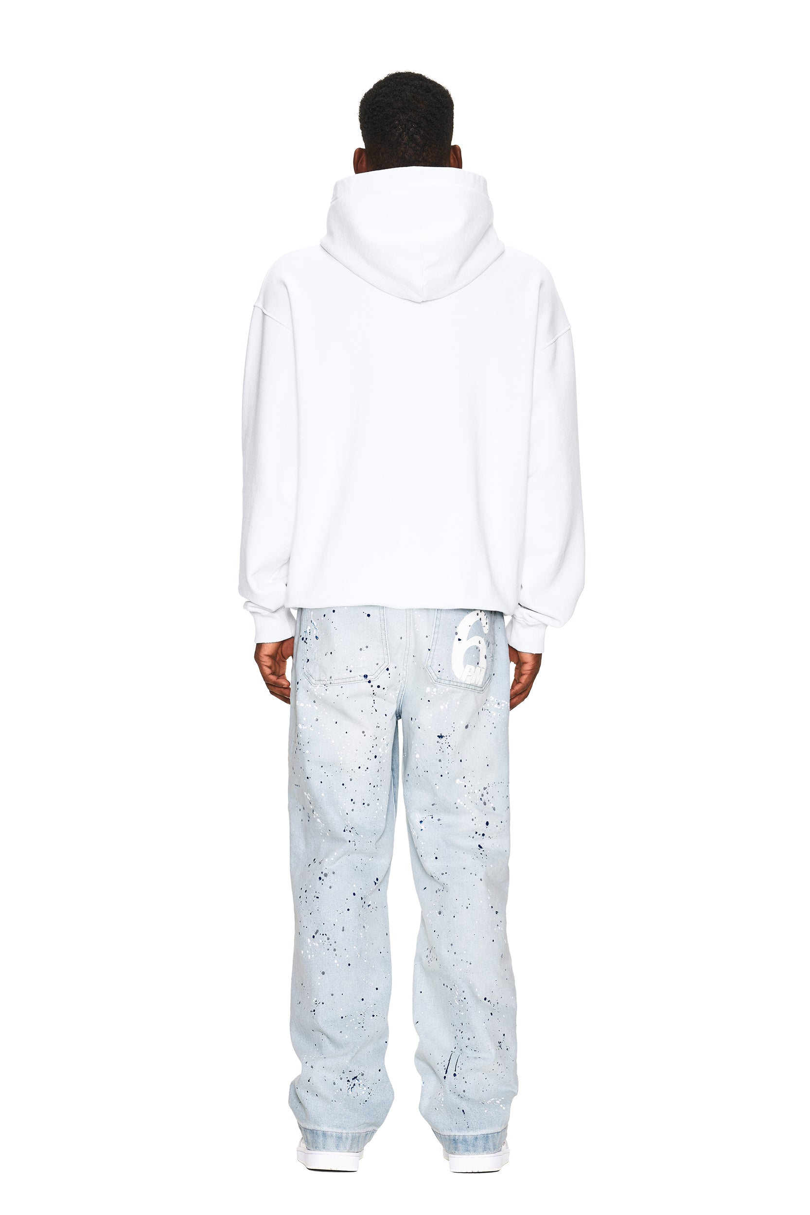 A person stands with their back to the camera, wearing the 6PM LOGO ZIP HOODIE in white and light blue paint-splattered jeans featuring a white graphic on the back pocket, against a plain white background.