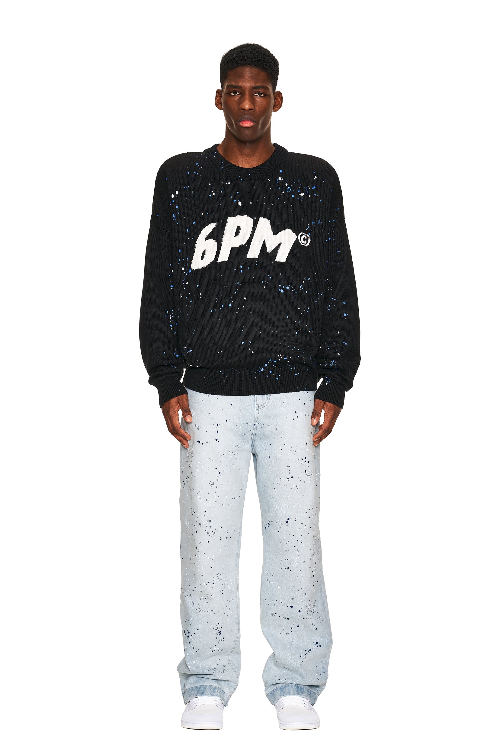 A man faces forward against a white background, wearing the 6PM LOGO KNIT BLACK sweater with bold white "6PM" lettering and blue-white splatter patterns, paired with matching light wash splatter jeans and white sneakers.
