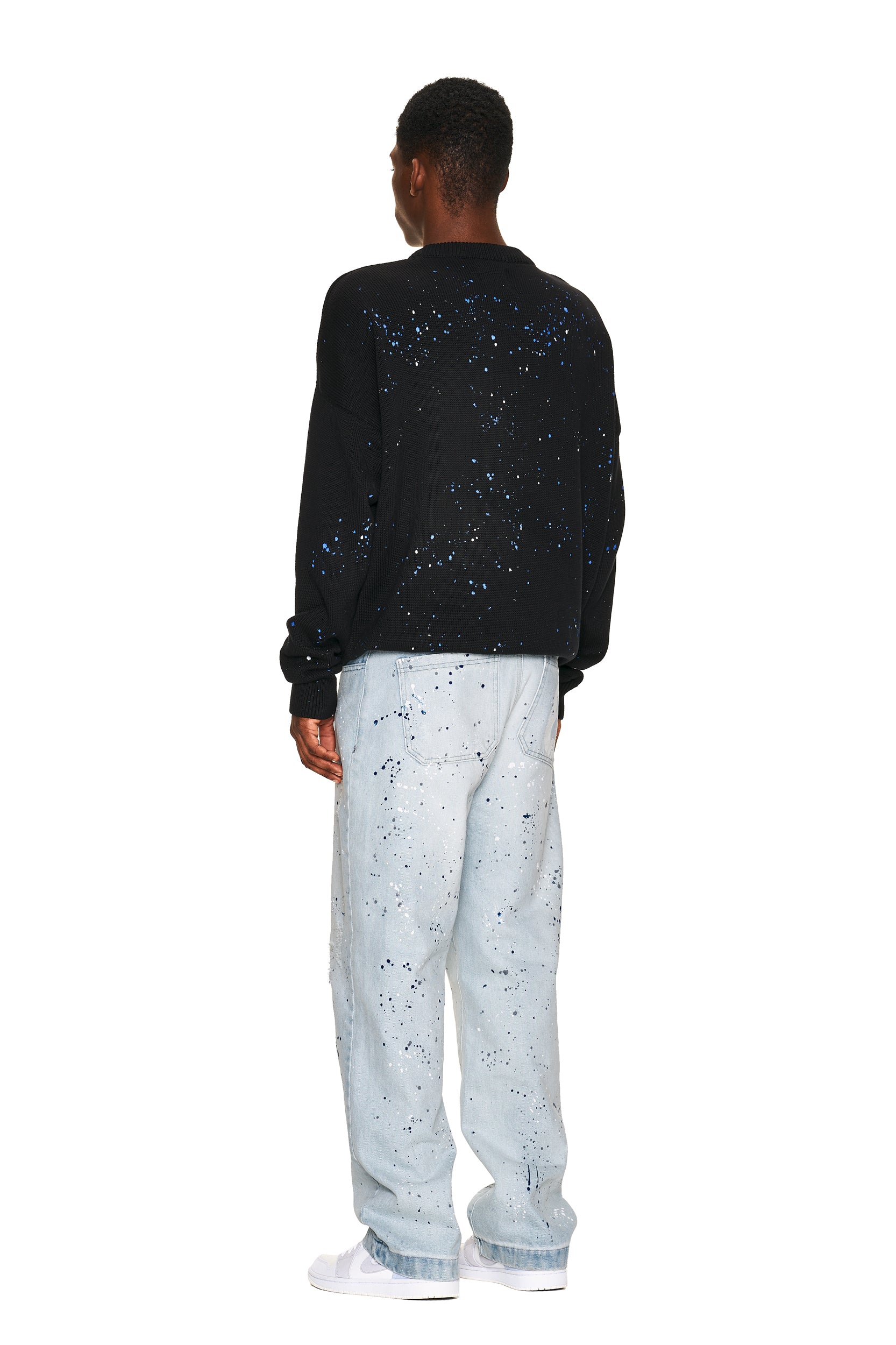 A person wears the 6PM LOGO KNIT BLACK sweatshirt with blue speckles, paired with loose light blue speckled jeans and white sneakers, standing against a plain white background.