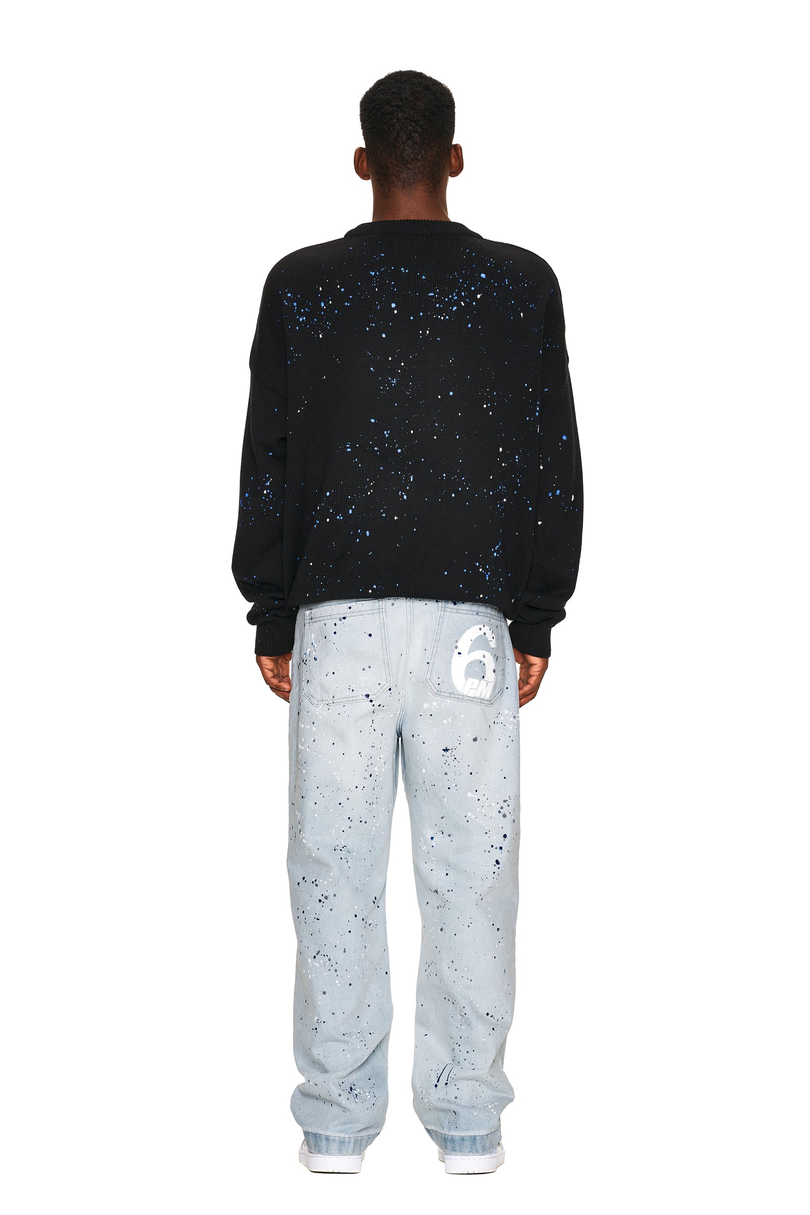 A person stands facing away against a white background, wearing the 6PM LOGO KNIT BLACK sweater with blue and white speckles and light blue jeans featuring paint splatters and a white “6” on the back pocket.