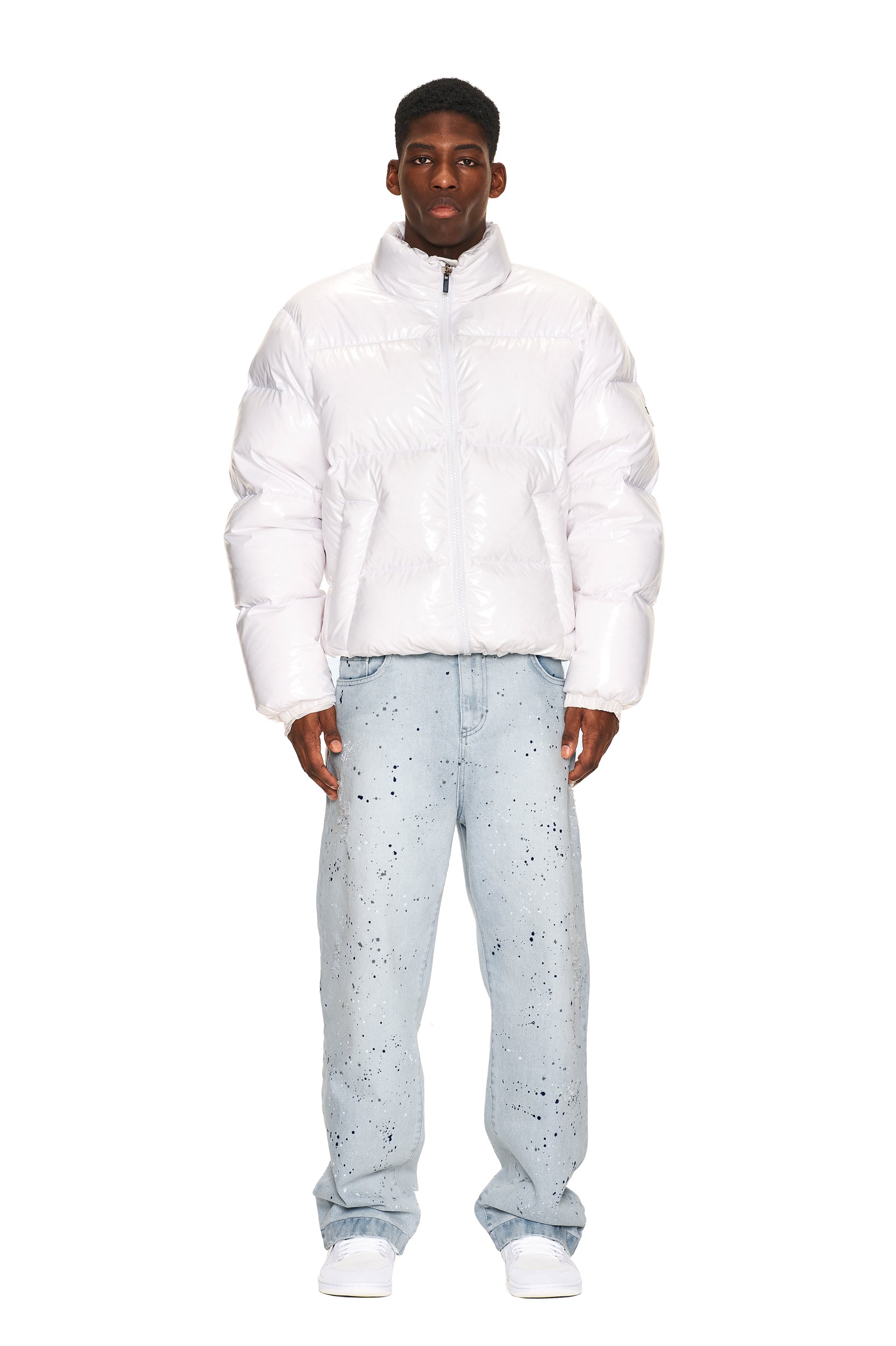 A man faces forward against a plain white background, wearing the 6PM DOWN JACKET WHITE SHINY, paired with light blue paint-splattered jeans and white sneakers.