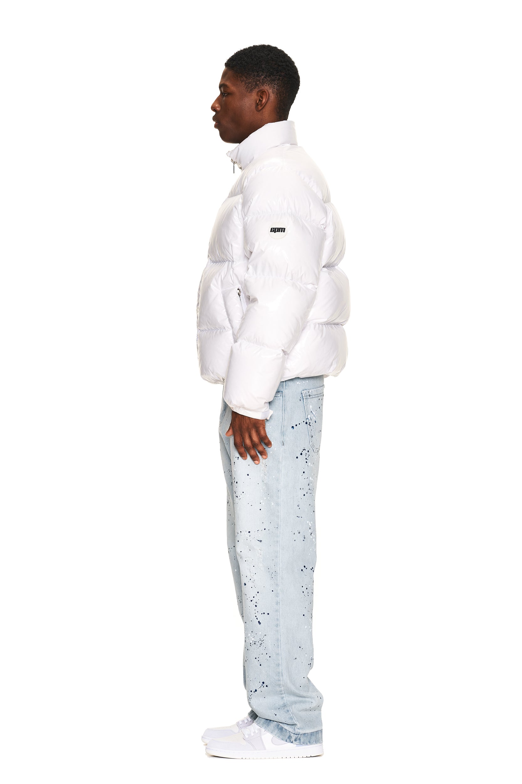 A man stands in profile against a white background, wearing the 6PM DOWN JACKET WHITE SHINY, light blue jeans with black splatter patterns, and white sneakers.