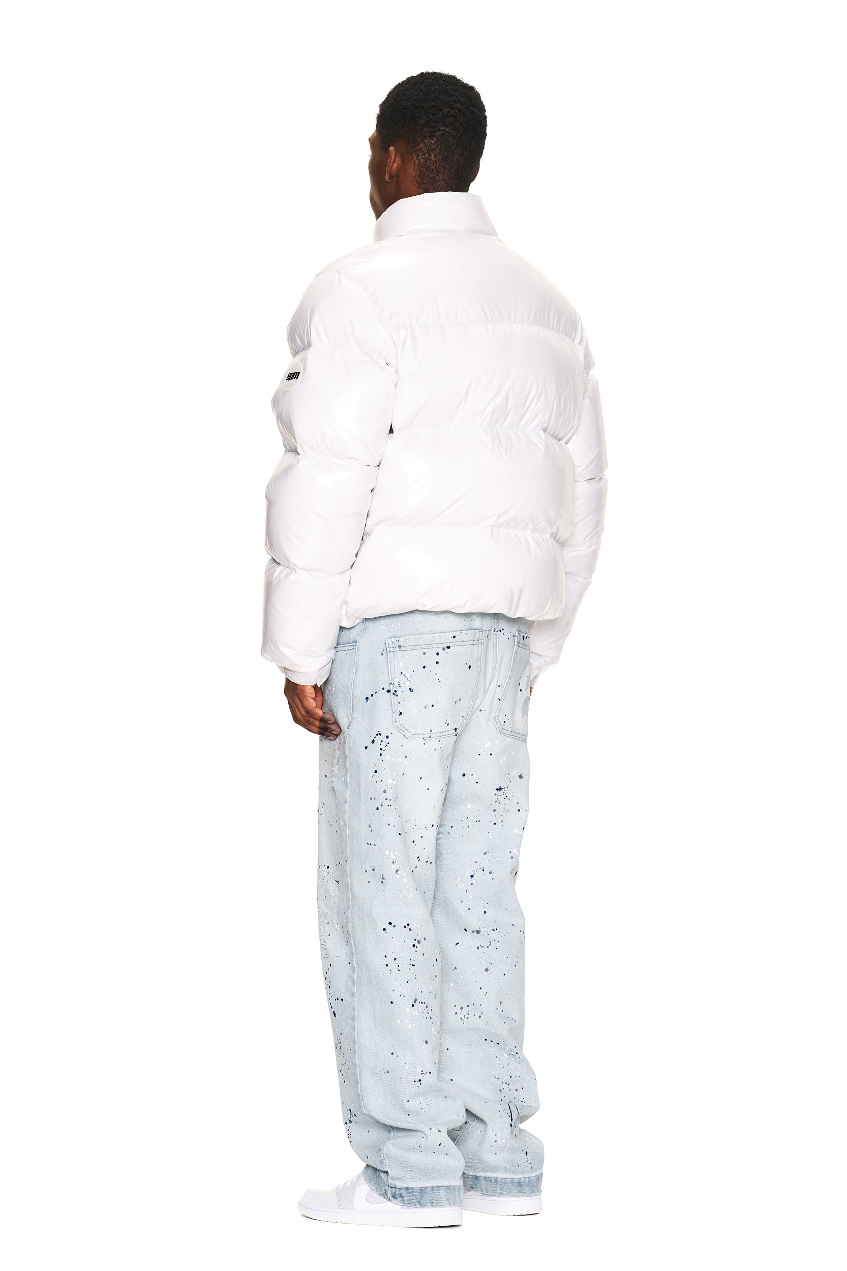A person faces away from the camera, wearing the 6PM DOWN JACKET WHITE SHINY, light blue paint-splattered jeans, and white sneakers against a white background.