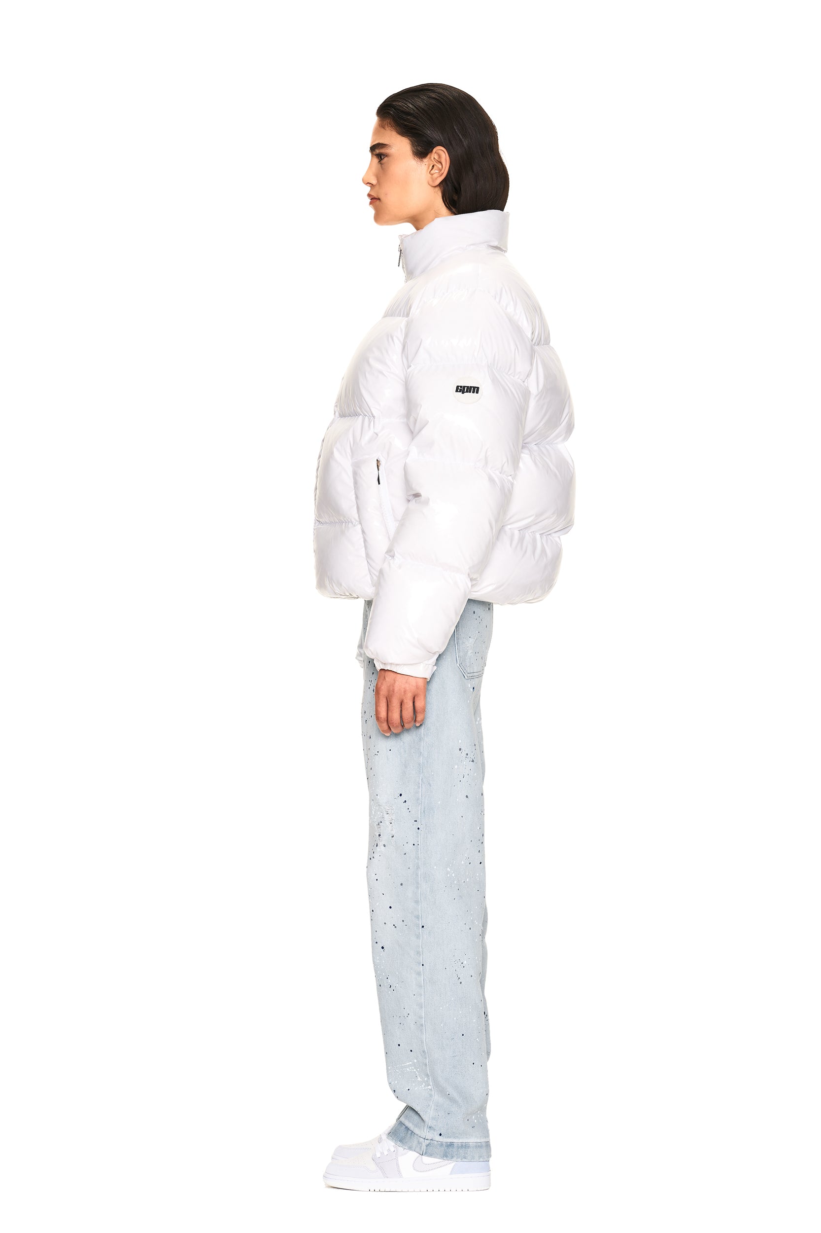 A person stands in profile against a plain white background, wearing the 6PM DOWN JACKET WHITE SHINY, light blue speckled jeans, and white sneakers.