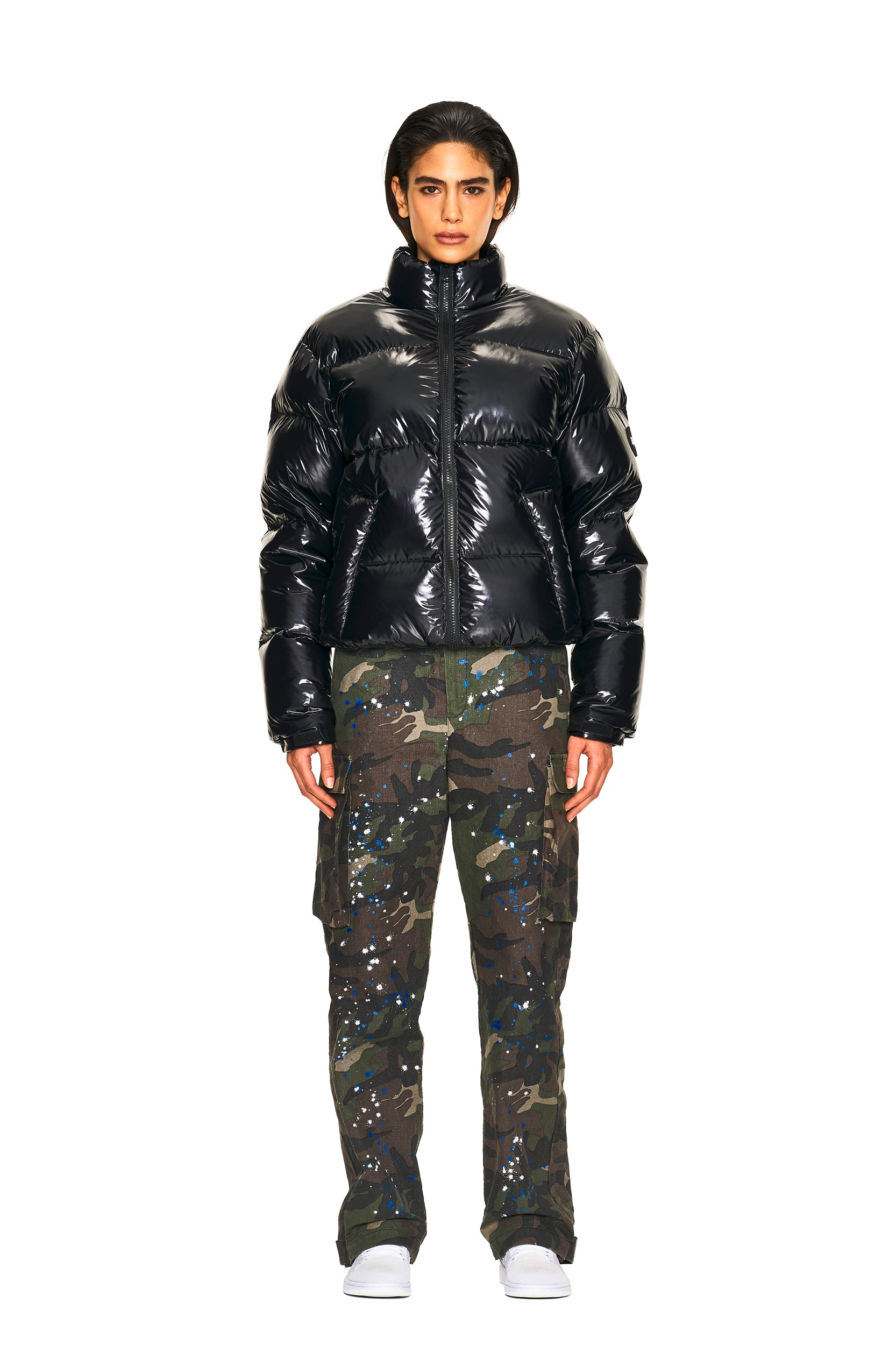 A person faces forward against a plain white background, wearing 6PM's CARGO PANTS CAMO with blue and white splatters, a shiny black puffer jacket, and white sneakers.