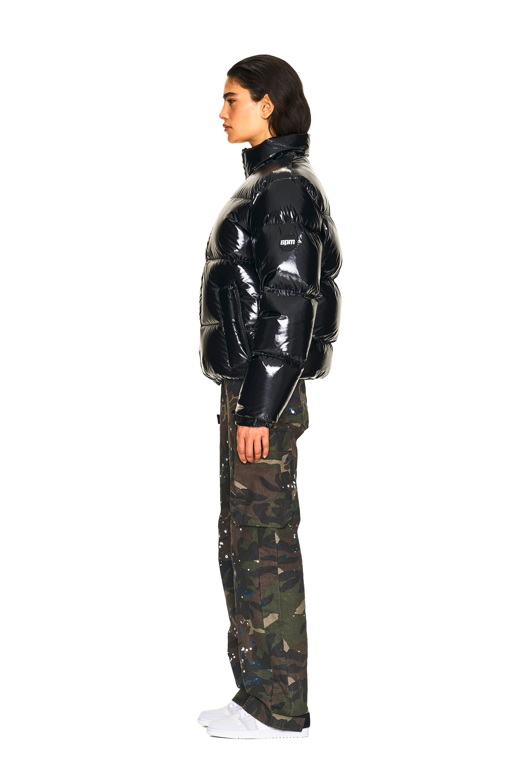 A person in profile wears a shiny black puffer jacket, 6PM CARGO PANTS CAMO with paint splatters, and white sneakers, posed against a plain white background.