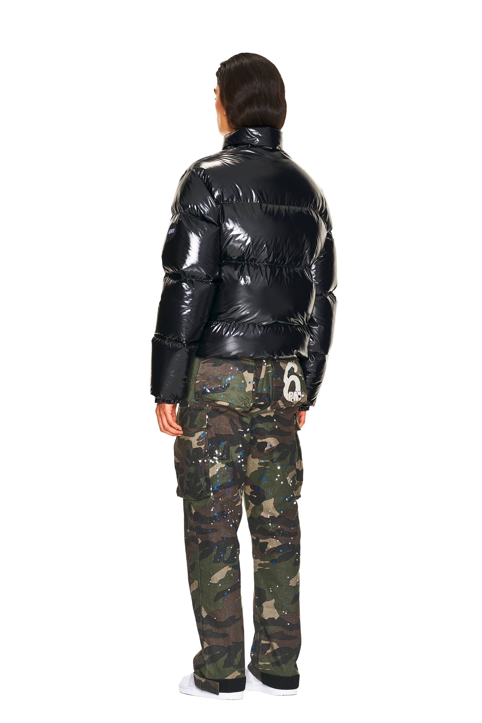 A person stands with their back to the camera, wearing a shiny black puffer jacket, 6PM CARGO PANTS CAMO with paint splatters, and white shoes.