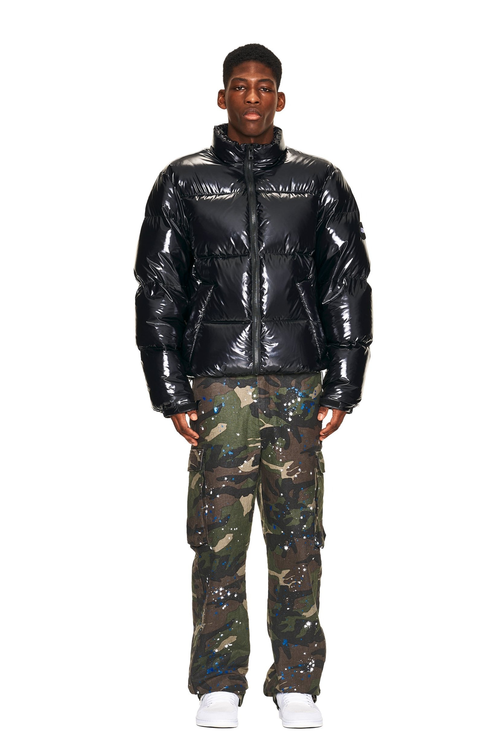 A person stands facing forward against a white background, wearing a shiny black puffer jacket, 6PM CARGO PANTS CAMO with blue paint splatters, and white shoes.