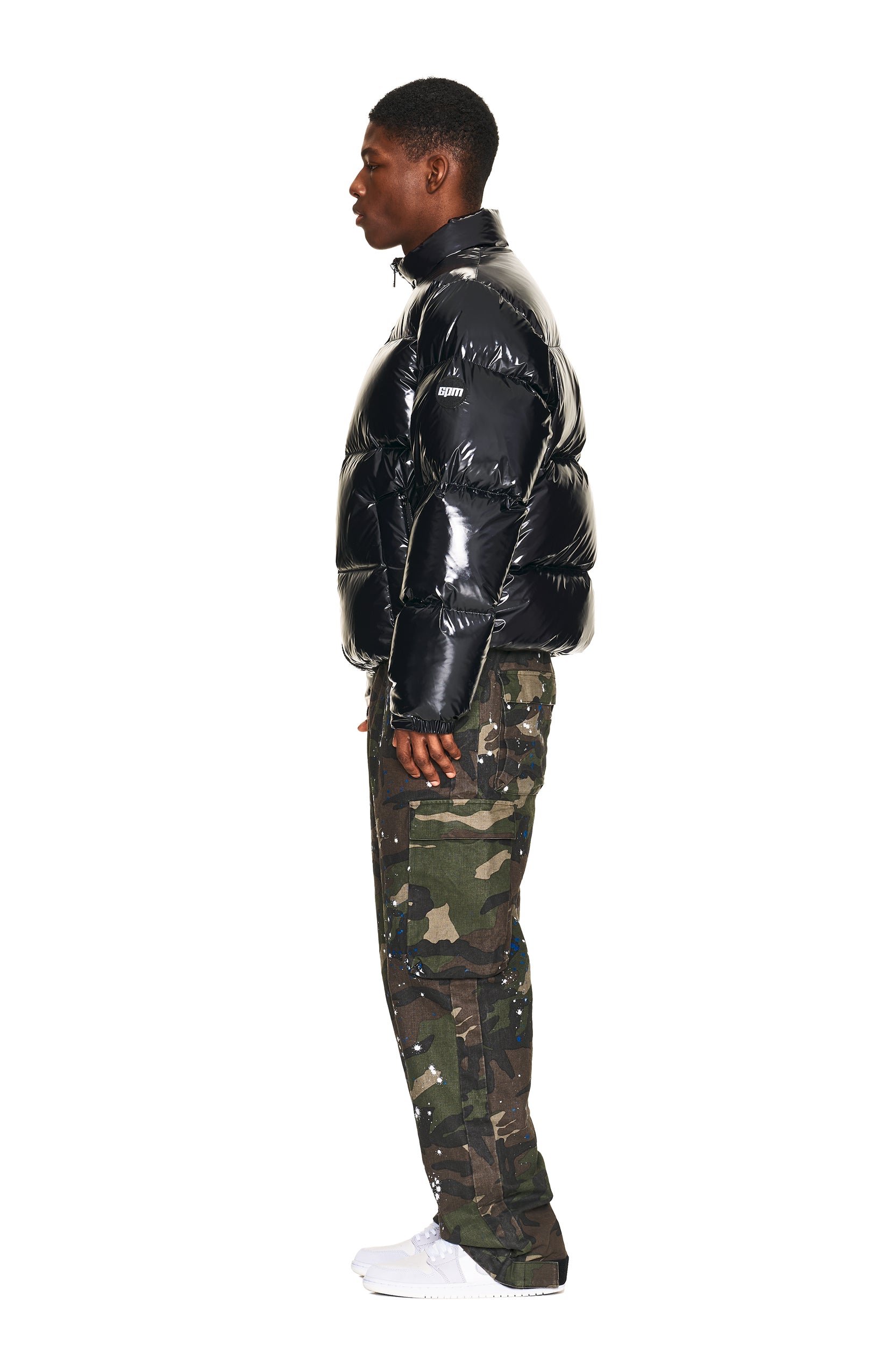 A man is shown in profile wearing a shiny black puffer jacket, 6PM CARGO PANTS CAMO, and white sneakers against a plain white background.