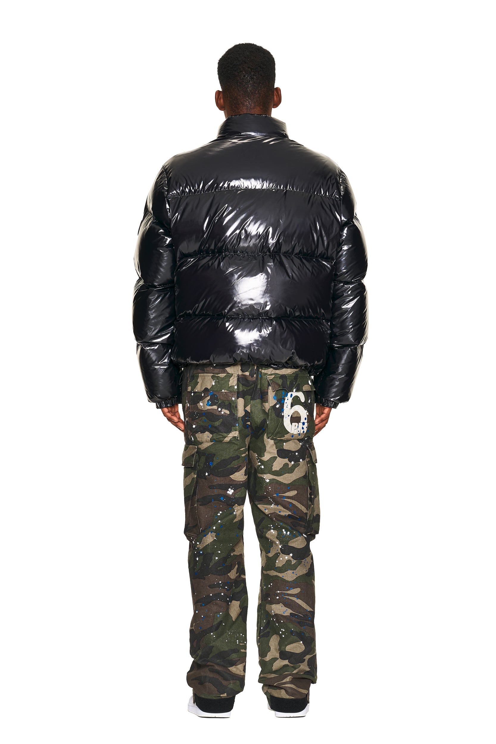 A person faces away against a white background, wearing a shiny black puffer jacket and 6PM CARGO PANTS CAMO featuring green camouflage, paint splatters, and a white 6 on the back pocket.