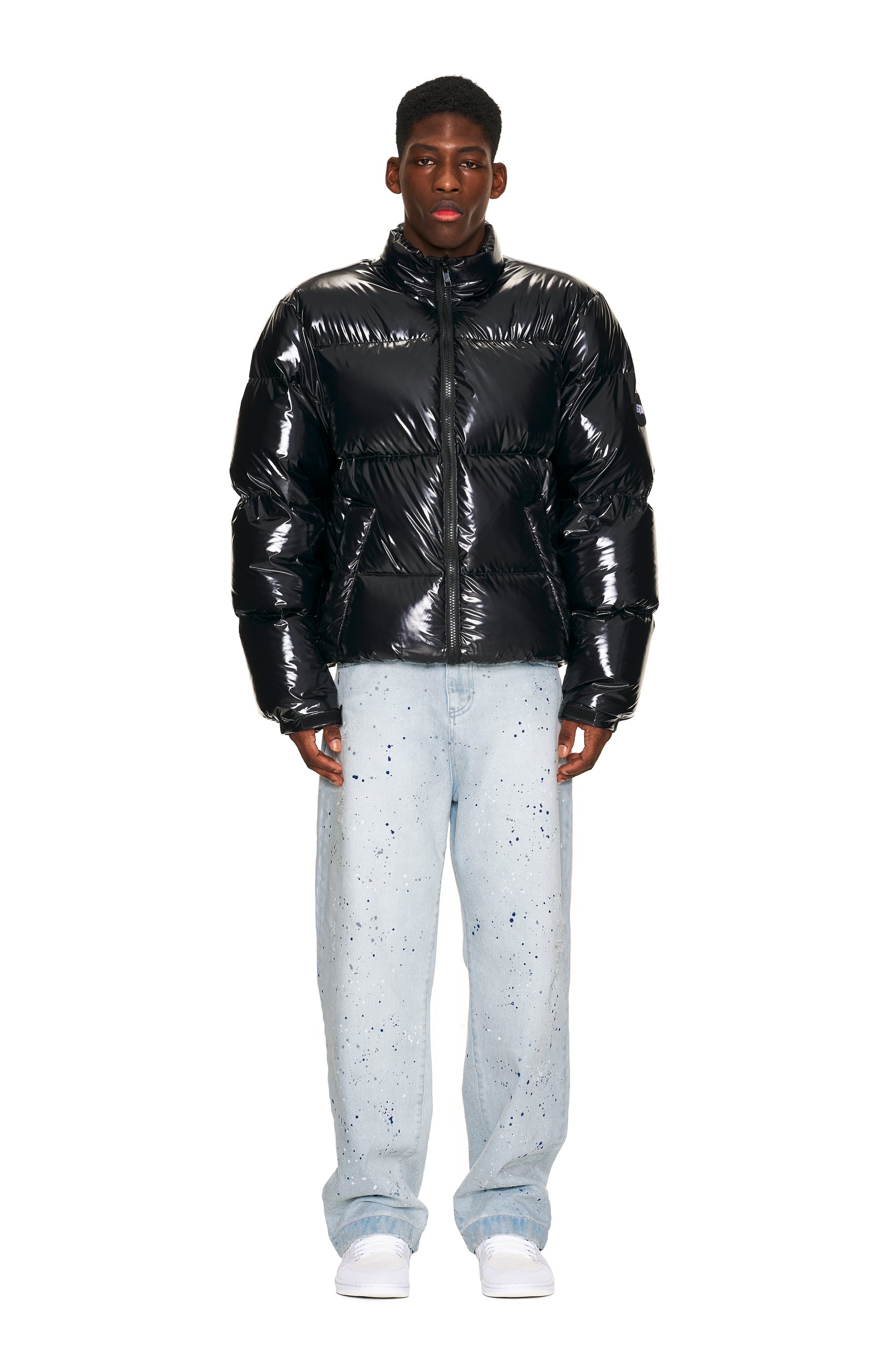 A man faces forward in a 6PM DOWN JACKET BLACK SHINY, light blue paint-splatter jeans, and white sneakers against a plain white background.
