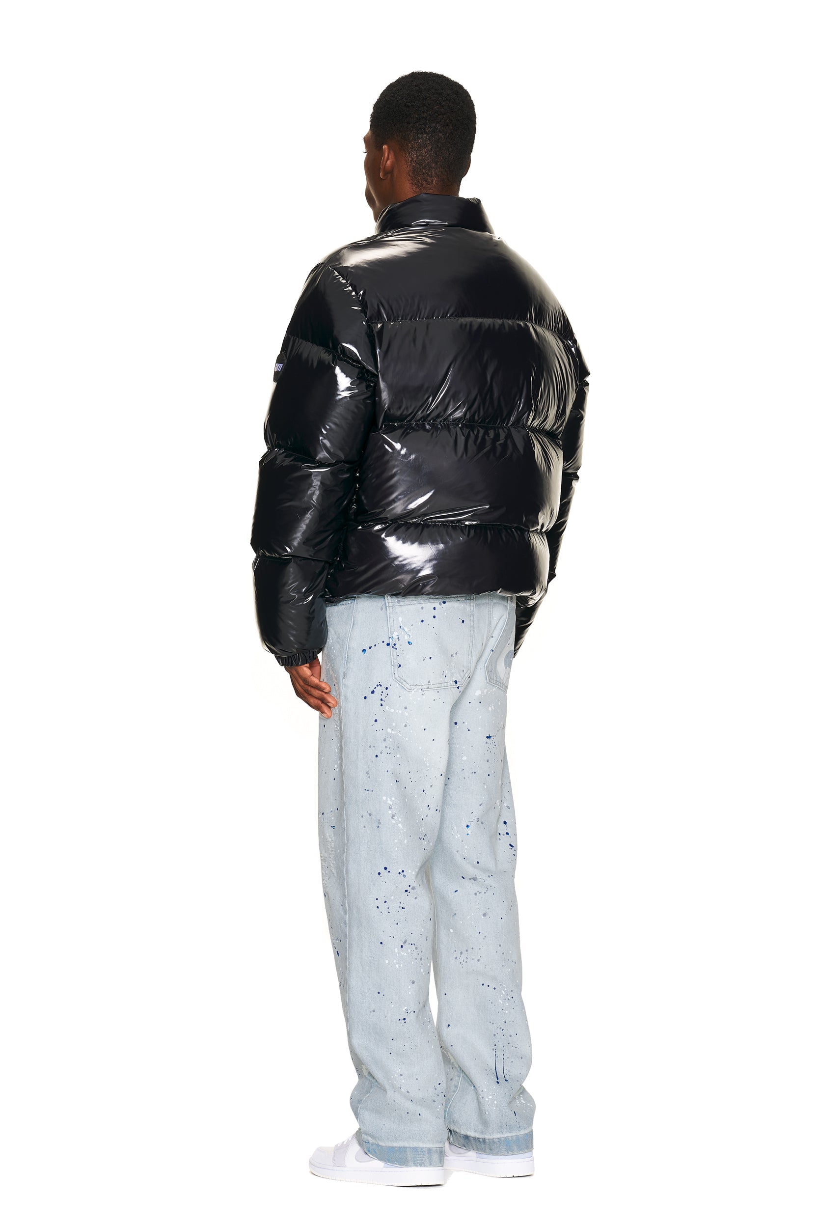 A person faces away, wearing the 6PM DOWN JACKET BLACK SHINY, light blue jeans with dark blue paint splatters, and white sneakers against a plain white background.