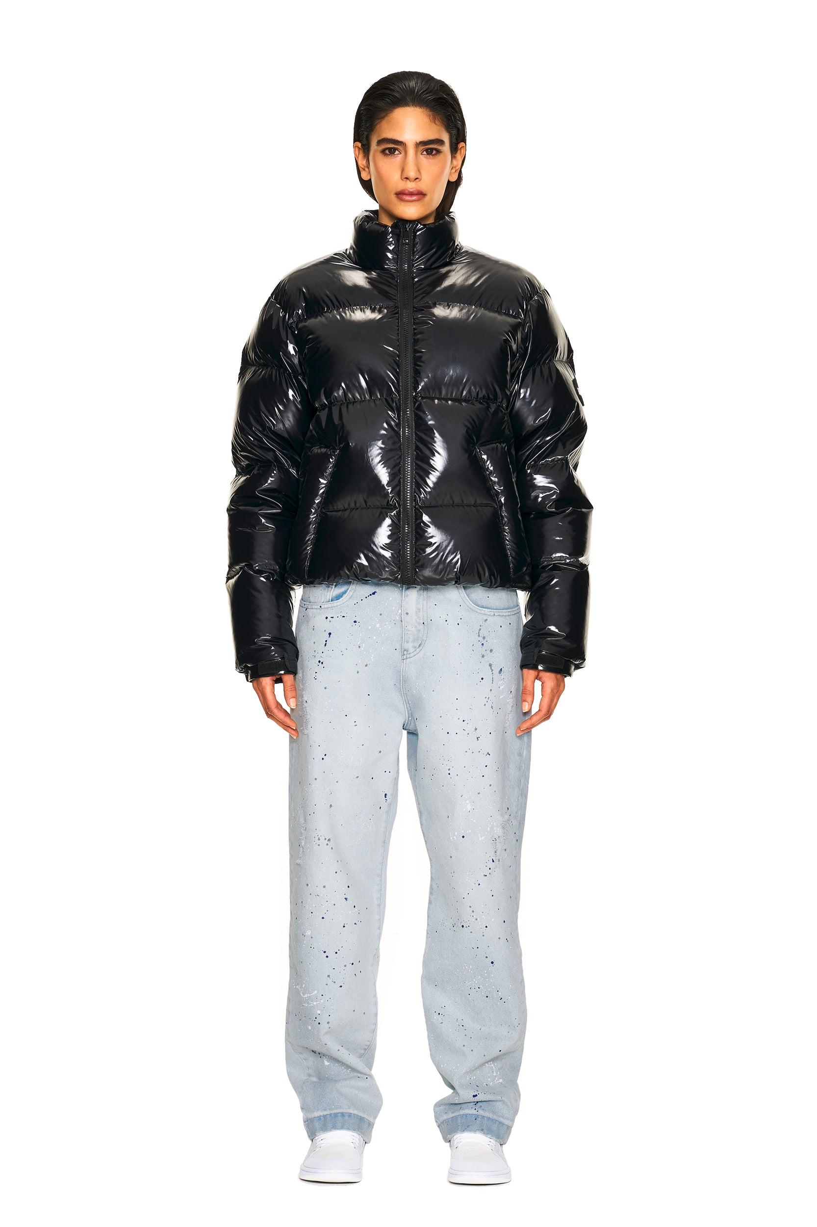 A person faces forward, wearing the 6PM DOWN JACKET BLACK SHINY, paired with loose light-wash splatter jeans and white slip-on shoes against a plain white background.