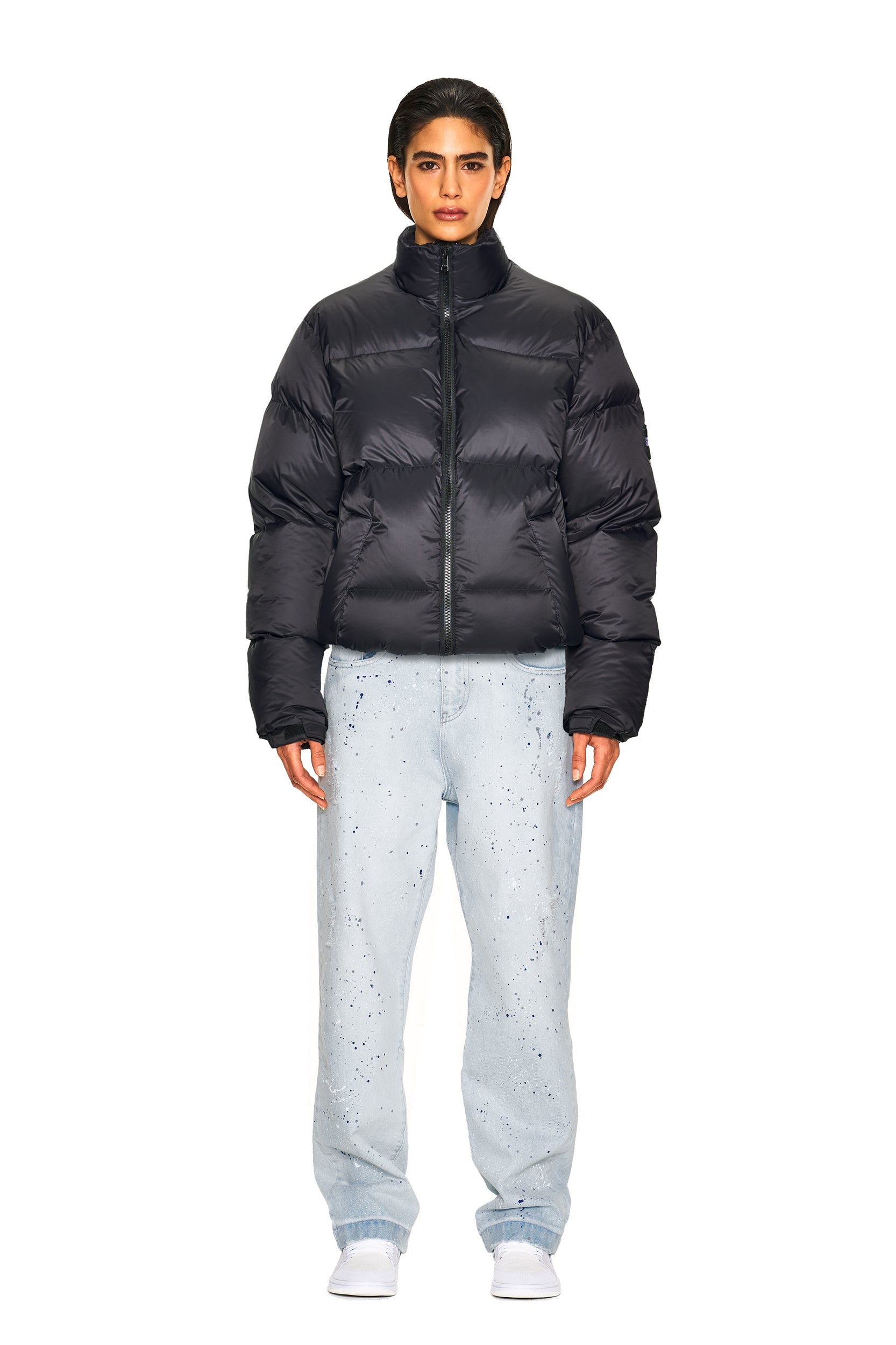 A person stands facing forward against a plain white background, wearing the 6PM DOWN JACKET BLACK MATTE, light blue paint-splattered jeans, and white shoes.