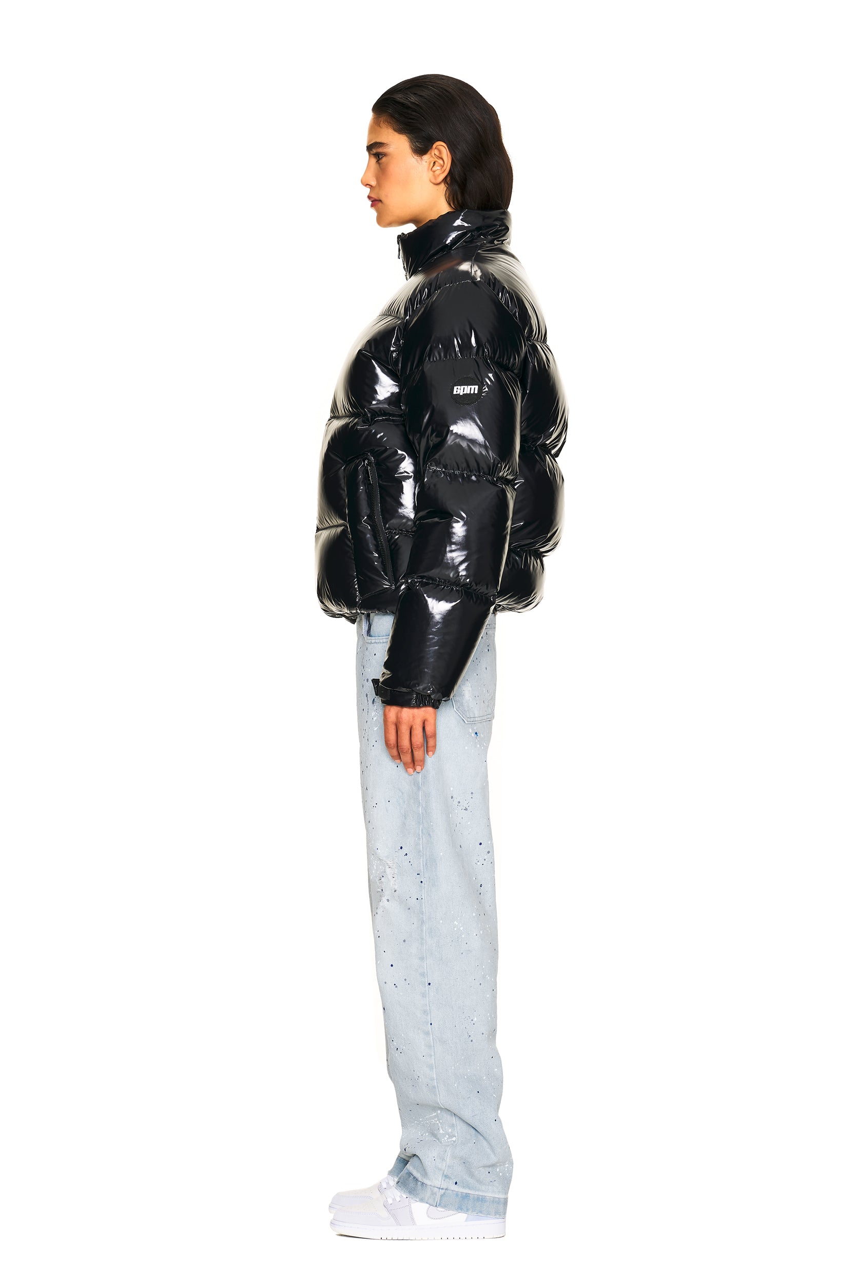 A person stands in profile wearing the 6PM DOWN JACKET BLACK SHINY, paired with light blue paint-splattered jeans and white sneakers, set against a plain white background.