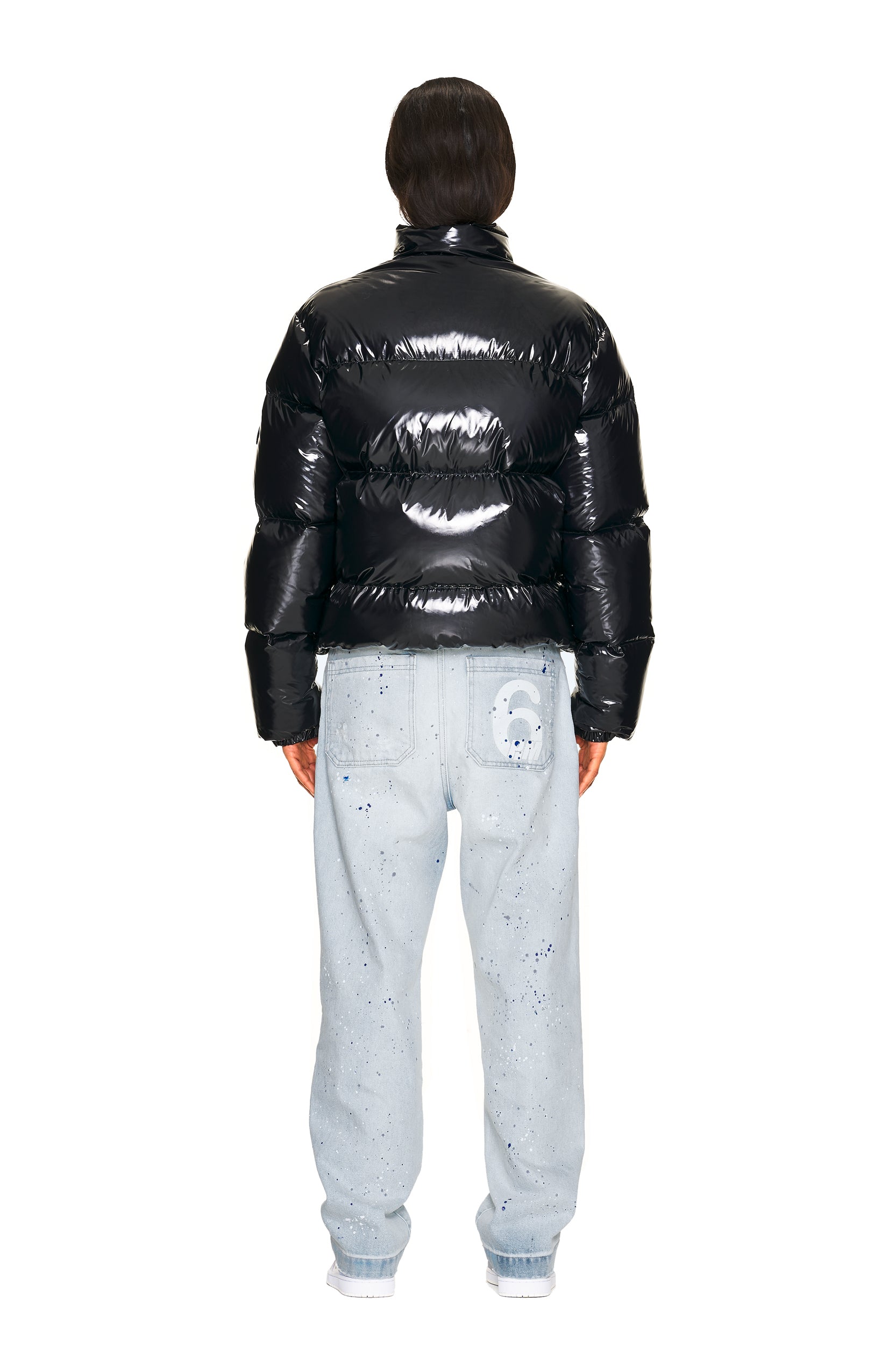 A person with short dark hair stands facing away, wearing the 6PM DOWN JACKET BLACK SHINY and light blue, slightly baggy jeans featuring a graphic on the back pocket, with their hands by their sides.