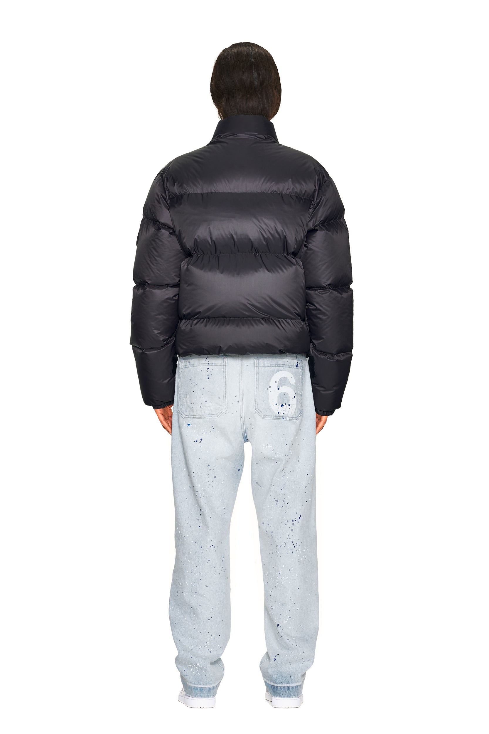 A person stands facing away, wearing the 6PM DOWN JACKET BLACK MATTE and light blue jeans with artistic paint splatters and a printed face on the back pocket, set against a white background.