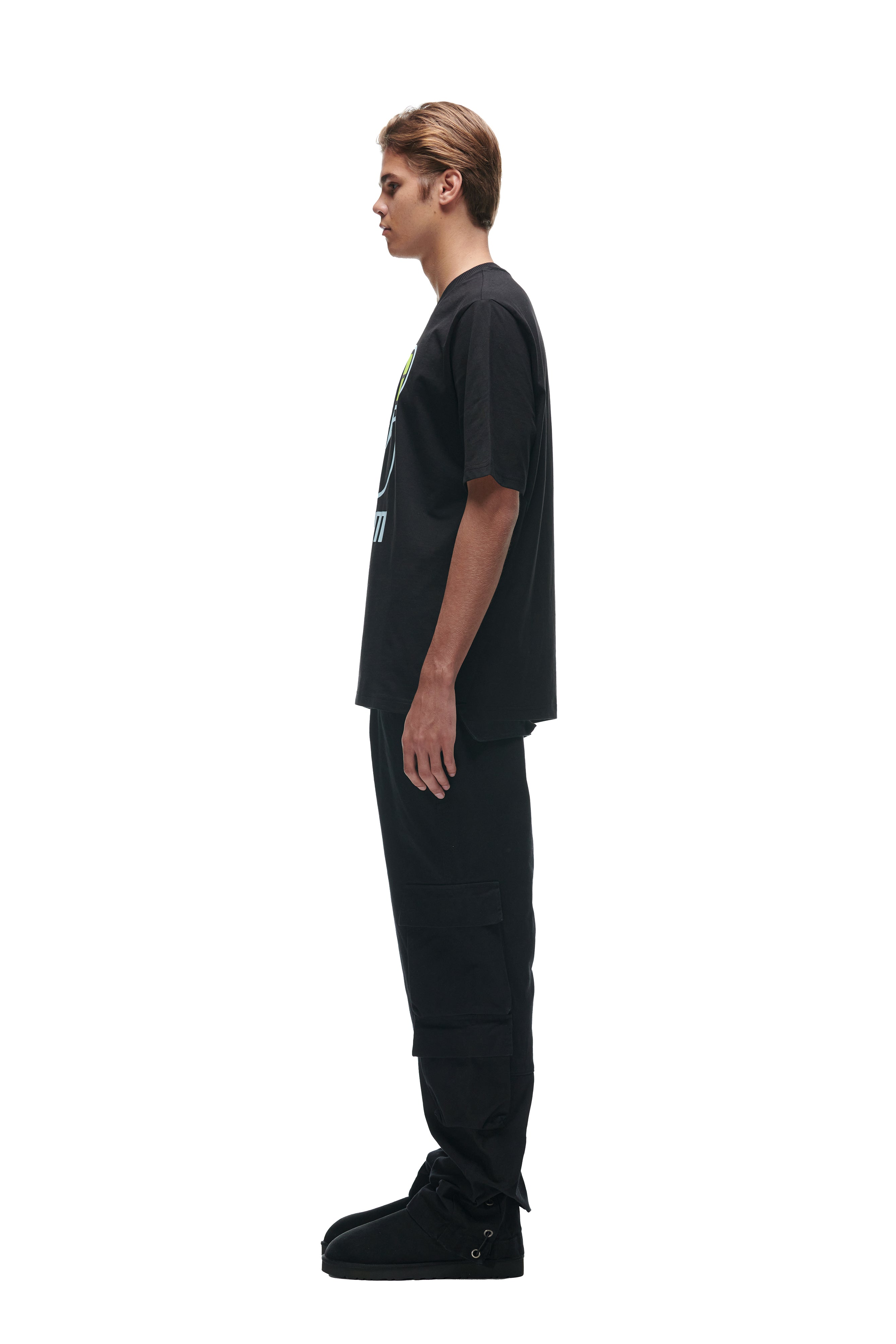 A young man stands in profile against a white background, wearing the 6PM FLOWER TSHIRT BLACK, paired with black cargo pants and boots. His short hair adds to the minimal look as his arms rest at his sides.