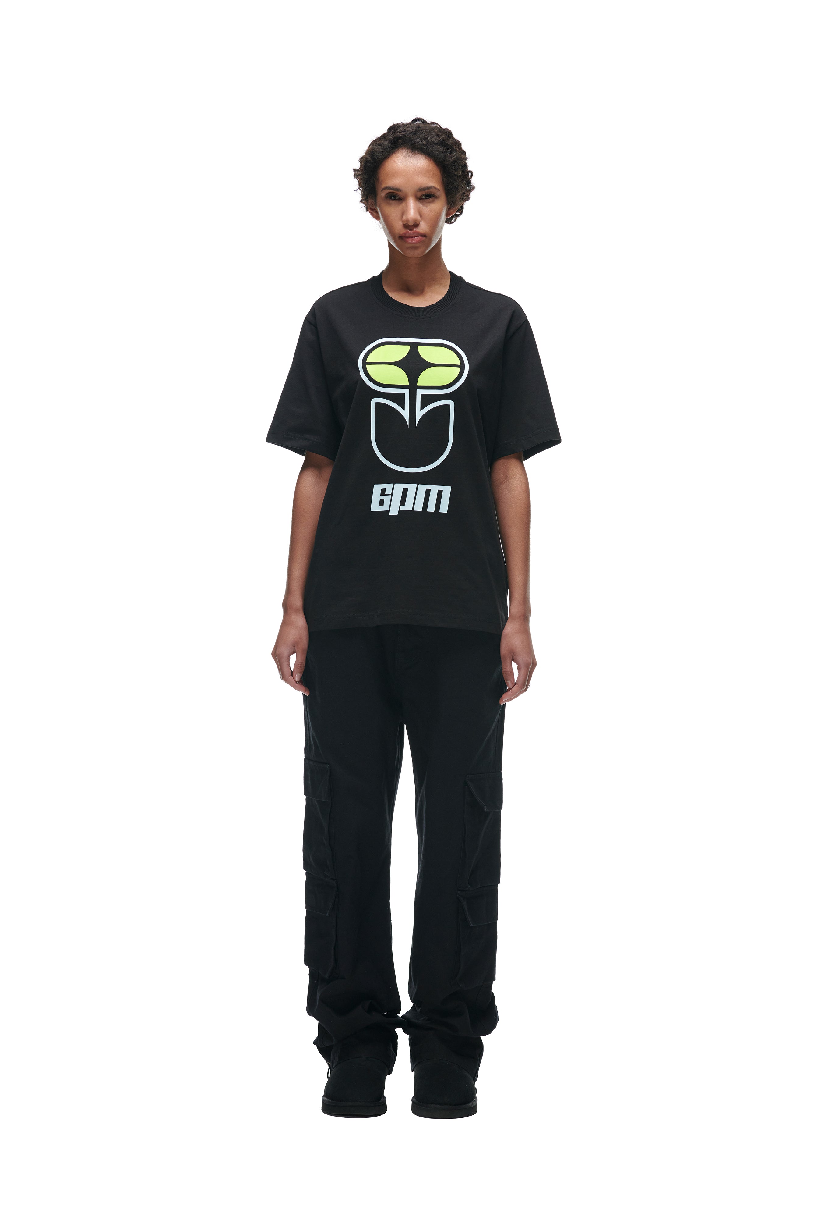 A person with short curly hair faces forward wearing the 6PM FLOWER TSHIRT BLACK, featuring a green and white graphic design, paired with black cargo pants and black shoes against a white background.