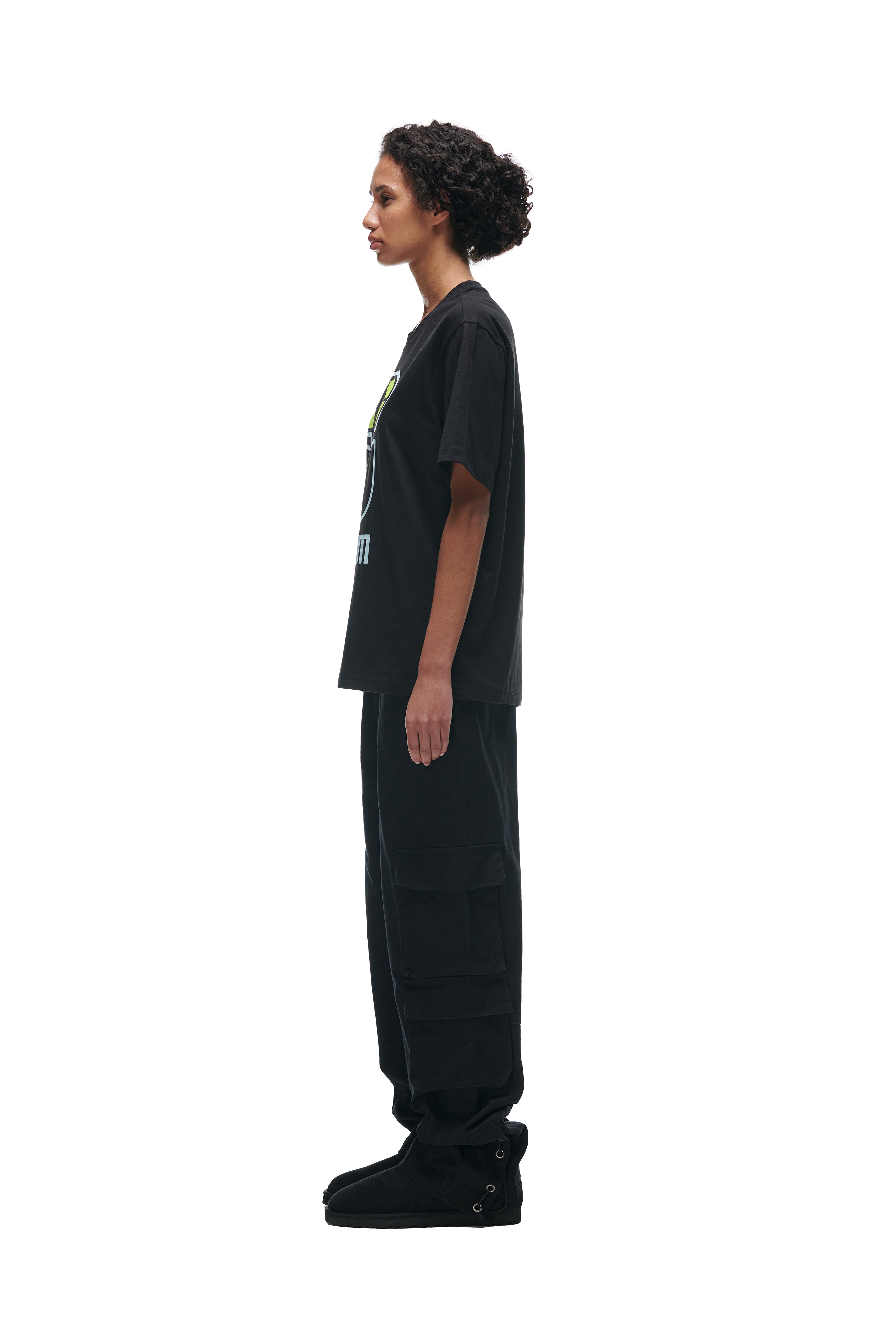 A person with short curly hair stands in profile against a white background, wearing the 6PM FLOWER TSHIRT BLACK, paired with black cargo pants and black shoes.