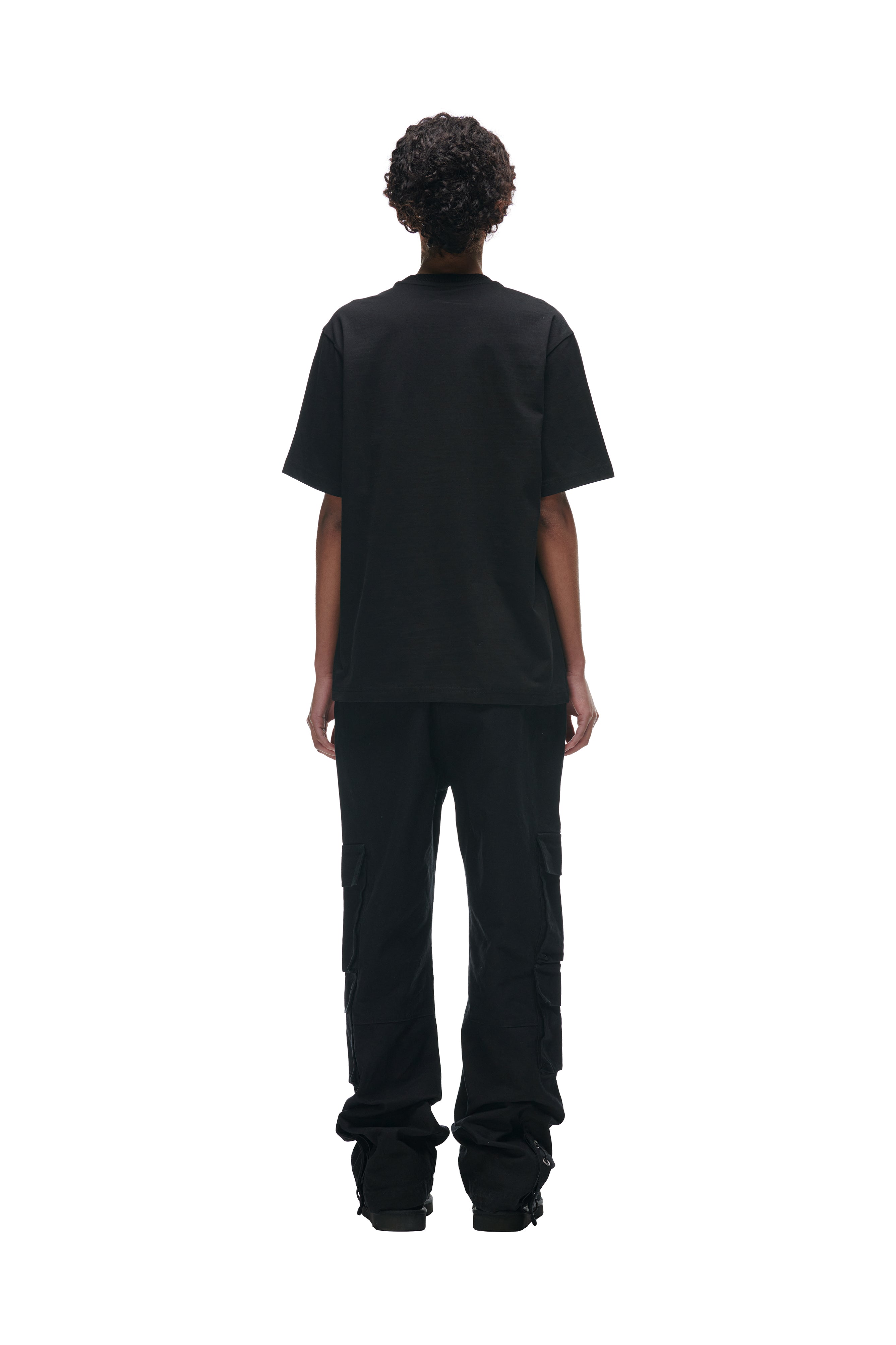A person with short curly hair stands facing away from the camera, wearing the 6PM FLOWER TSHIRT BLACK, black cargo pants, and black shoes against a plain white background.