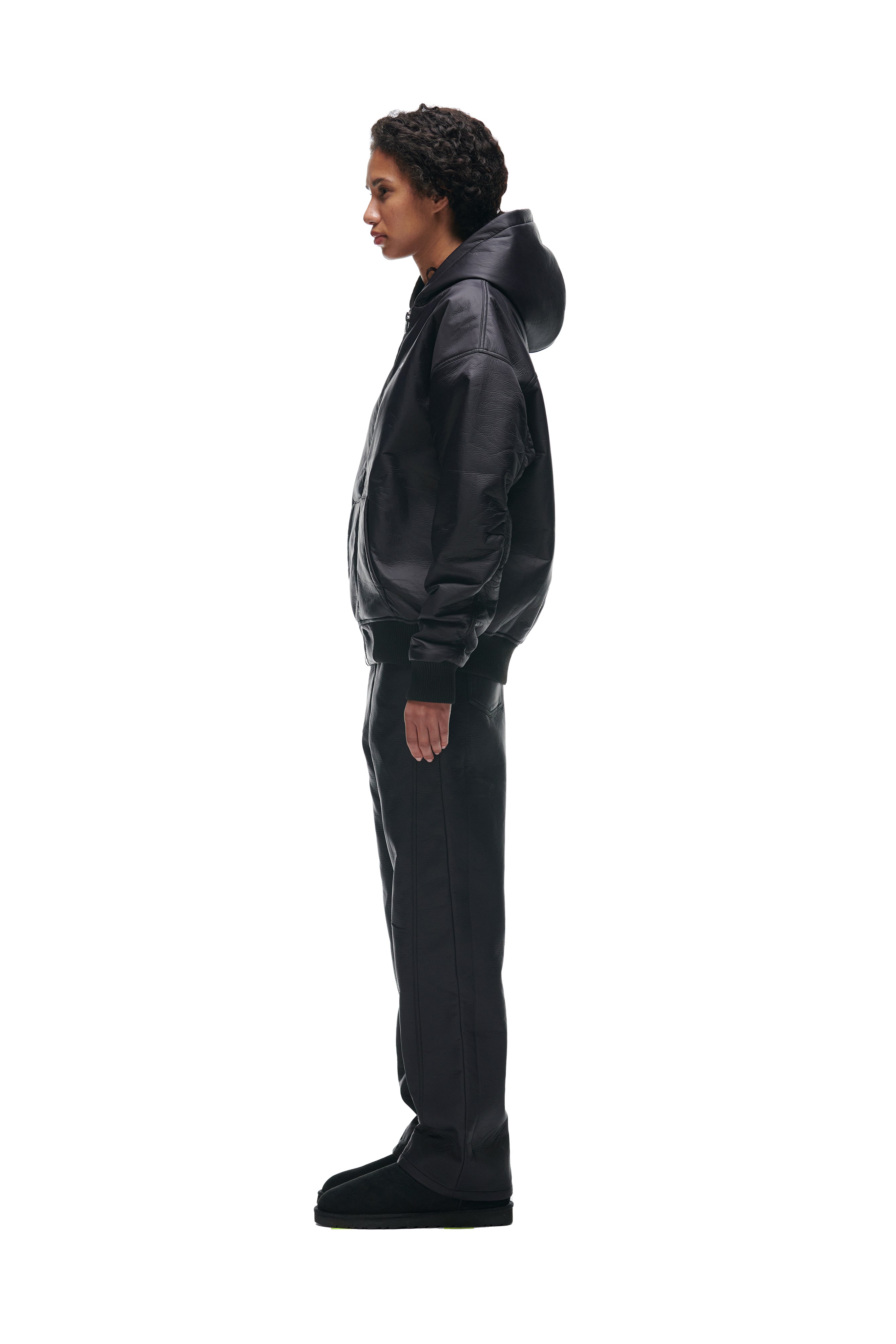 A person with short curly hair stands in profile against a plain white background, wearing the 6PM CRINKLE ZIP JACKET BLACK, paired with black pants and black shoes.