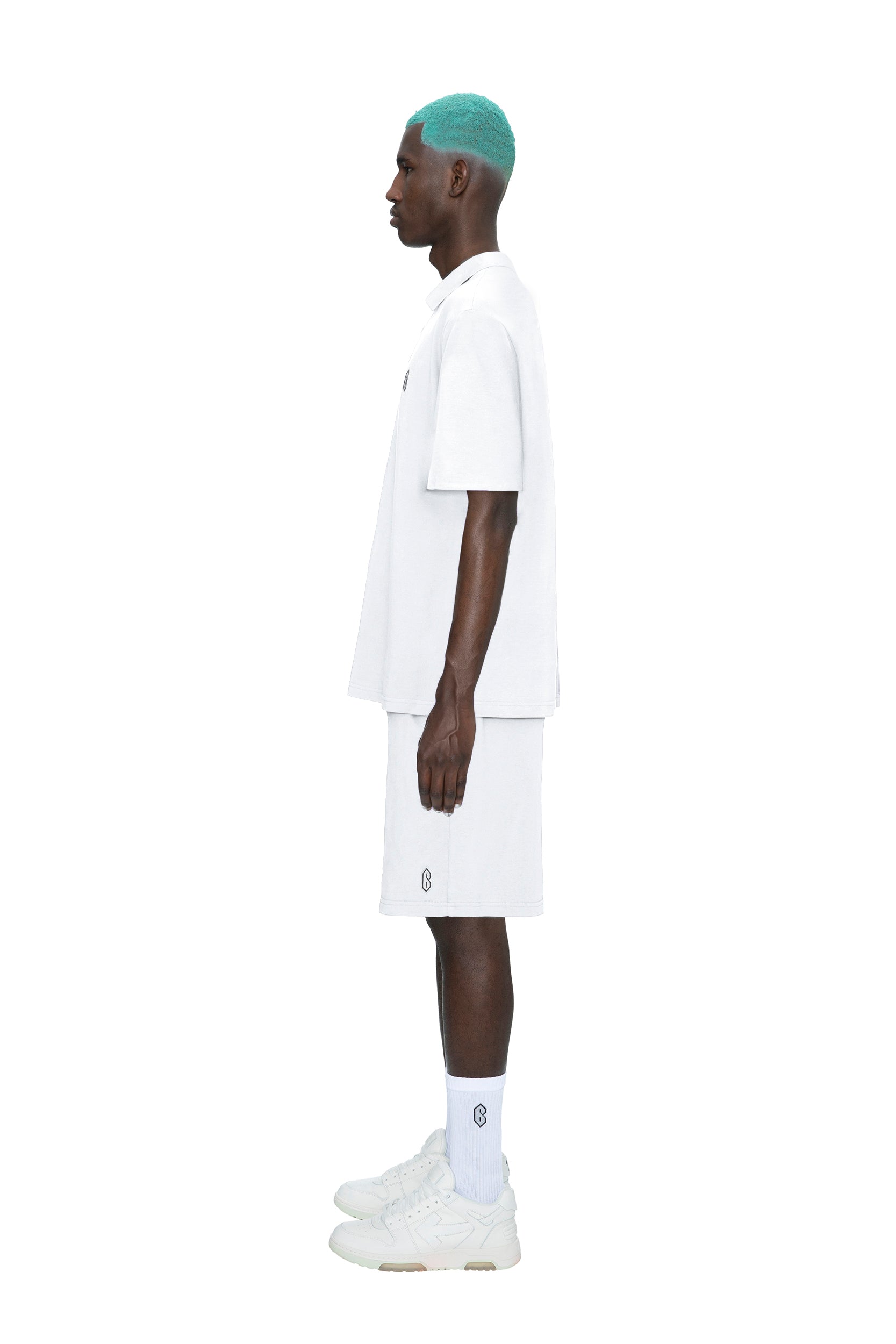 A person with teal hair stands in profile against a white background, wearing an oversized 6PM POLO SHIRT WHITE, white shorts, white socks, and white sneakers.