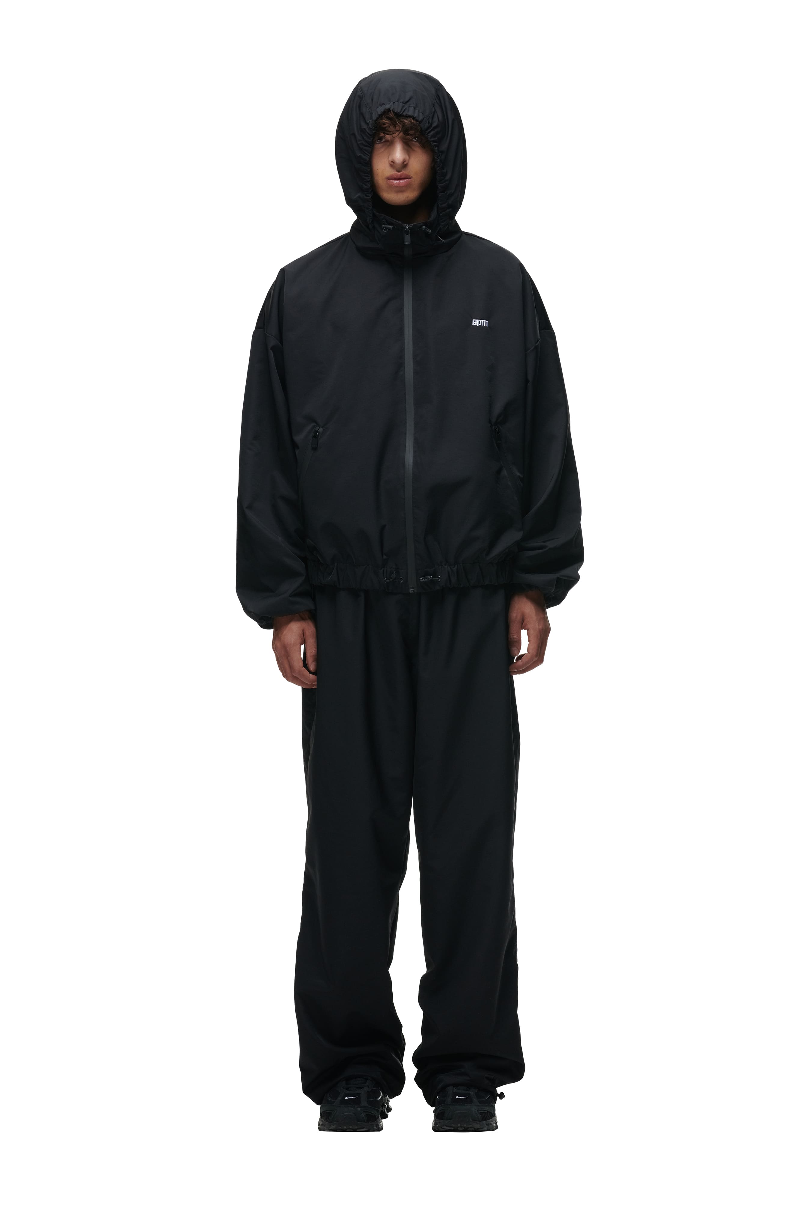 A person faces forward wearing the 6PM TRACK JACKET BLACK with loose black pants and black sneakers, set against a plain white background.