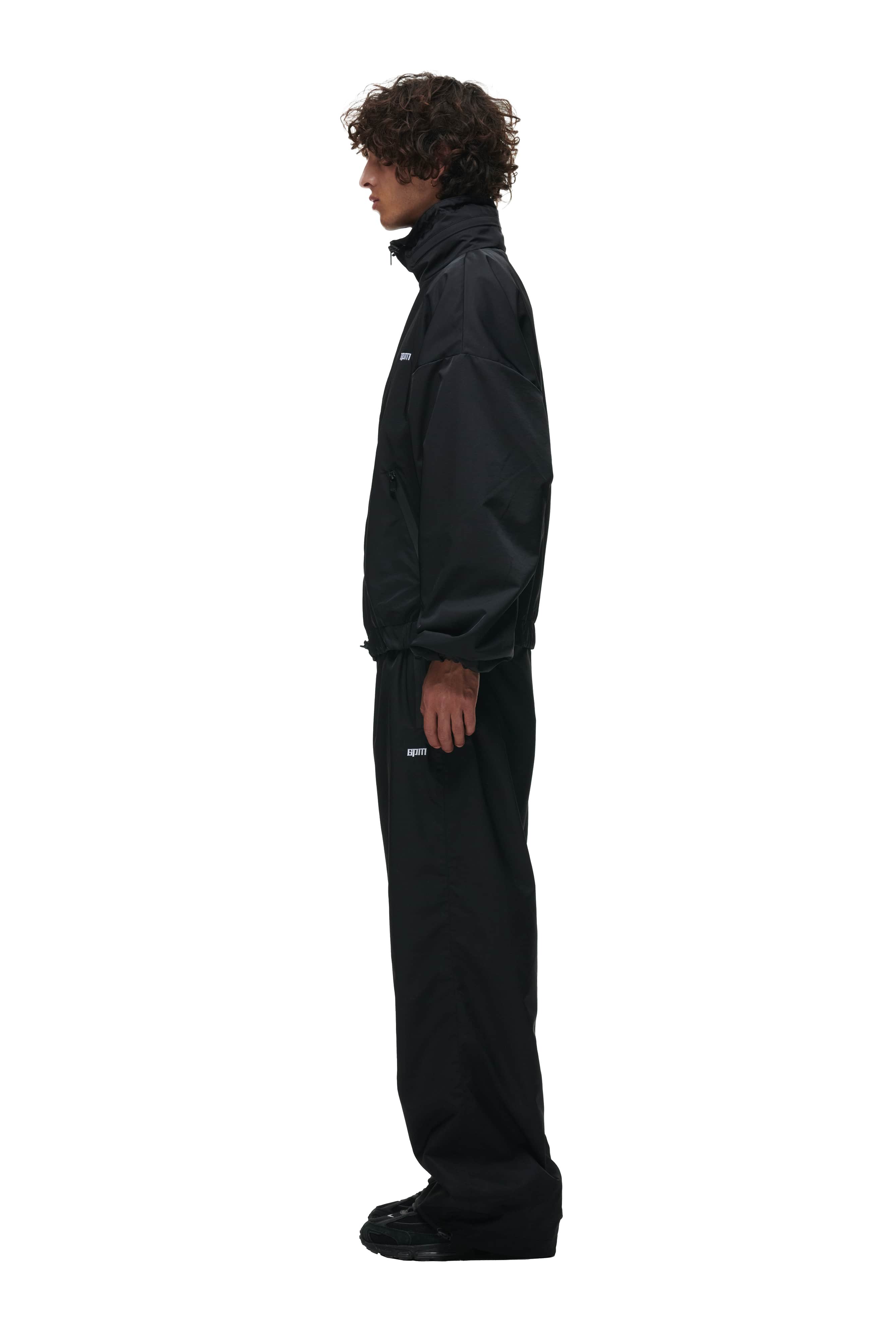 A person with curly hair stands in profile against a plain white background, wearing 6PM TRACK PANTS BLACK paired with a black zip-up jacket and black shoes.