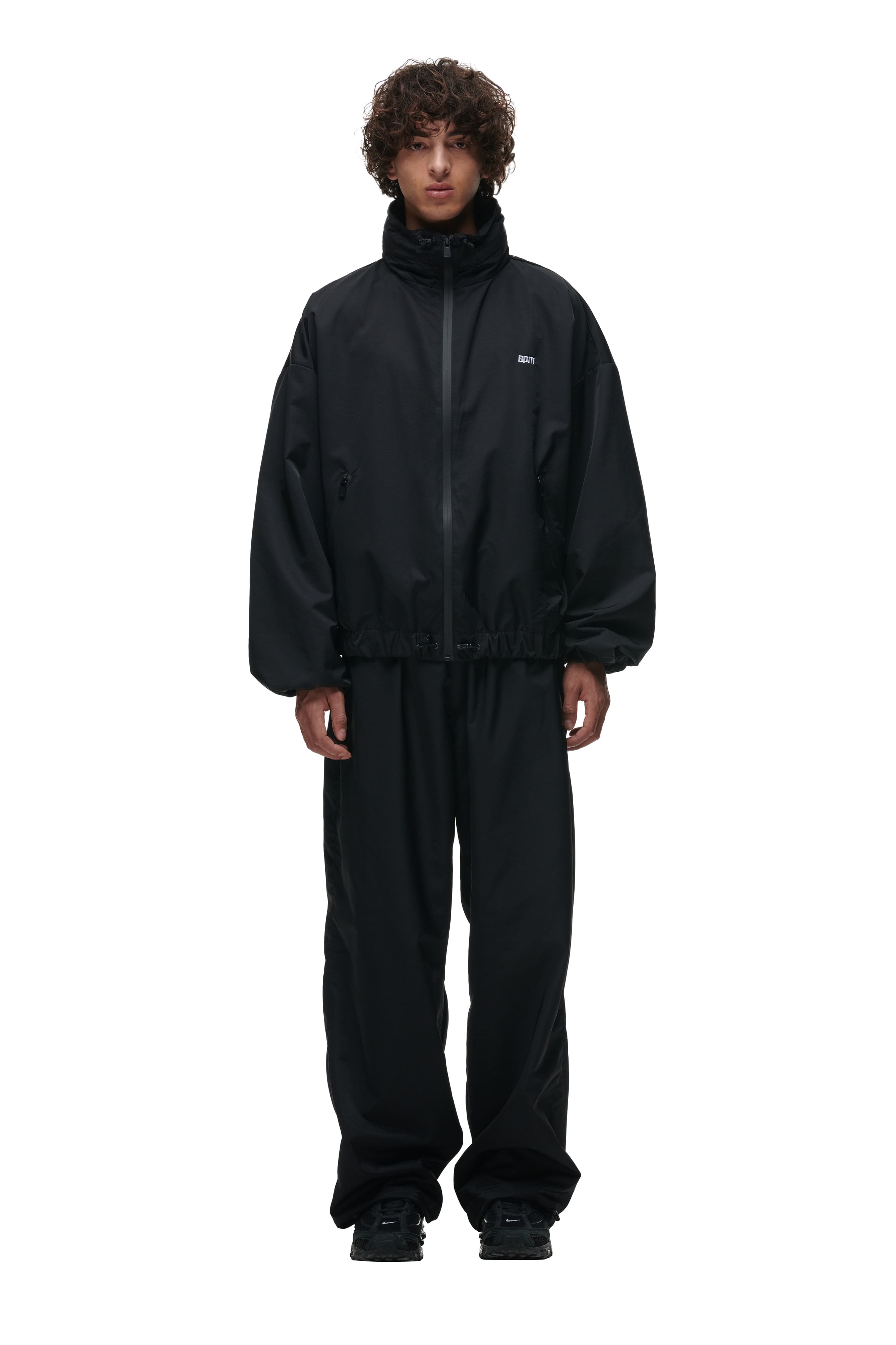 A person with curly hair stands facing forward against a plain white background, wearing 6PM TRACK PANTS BLACK paired with a loose-fitting, black zip-up jacket.