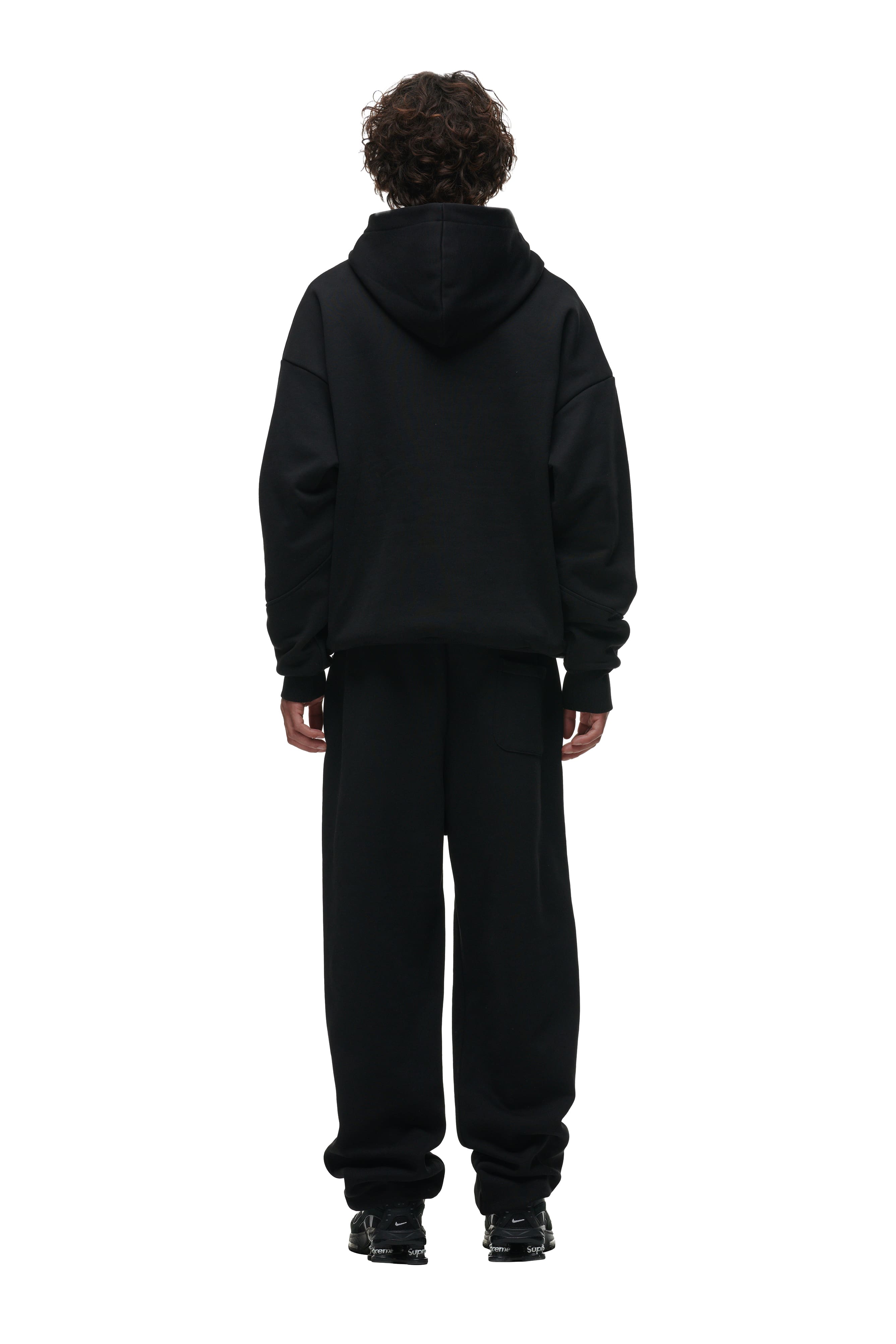 A person with curly hair is facing away from the camera, wearing the 6PM DOUBLE LAYER PLAY HOODIE BLACK, black sweatpants, and black shoes against a plain white background.