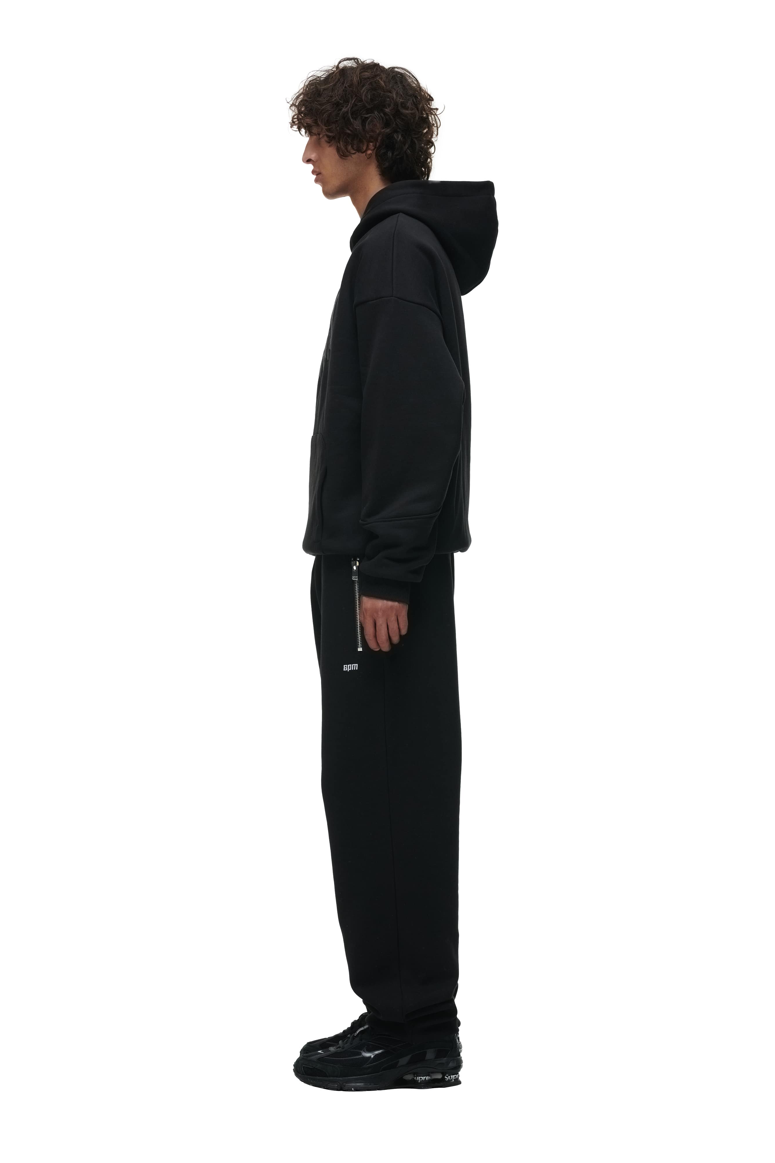 A person with curly hair stands in profile against a white background wearing the 6PM DOUBLE LAYER PLAY HOODIE BLACK, along with black sweatpants and black shoes.