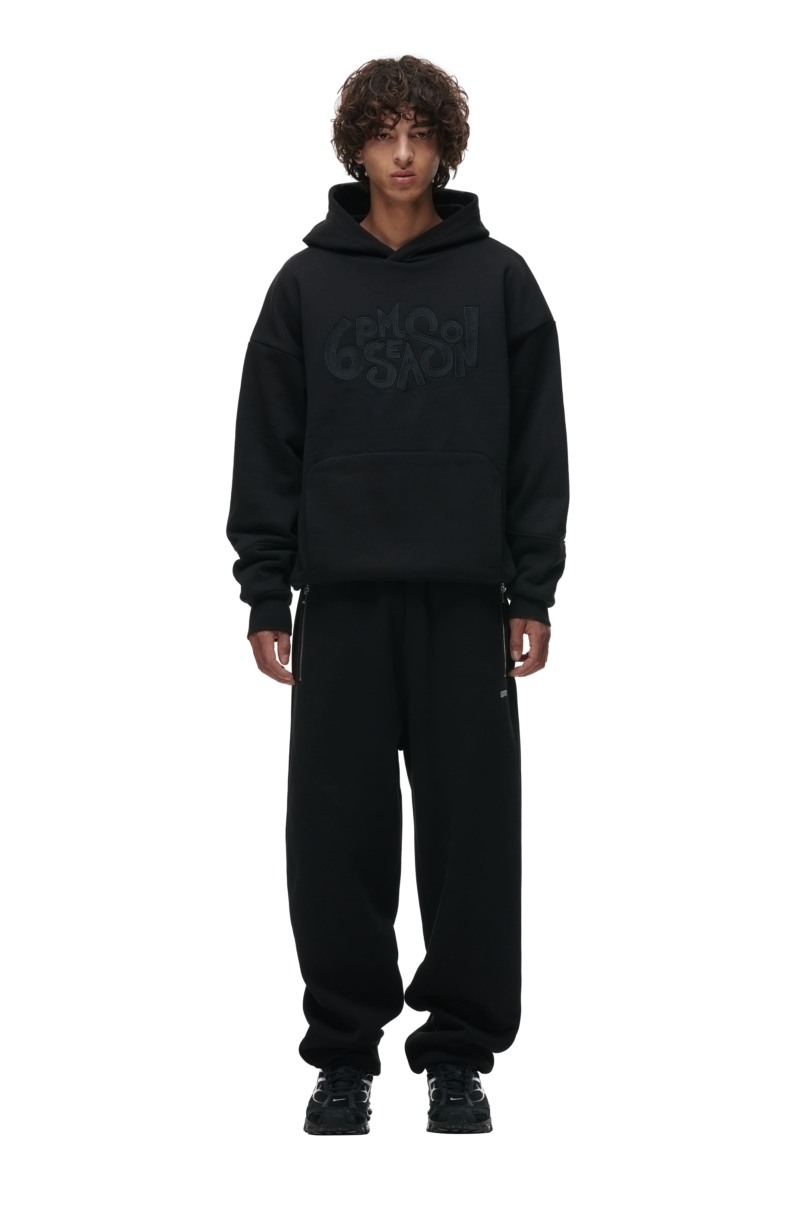 A person with curly hair wears the 6PM DOUBLE LAYER PLAY HOODIE BLACK, featuring a loose fit, large front pocket, and graphic text, paired with baggy black pants and black sneakers, standing against a white background.