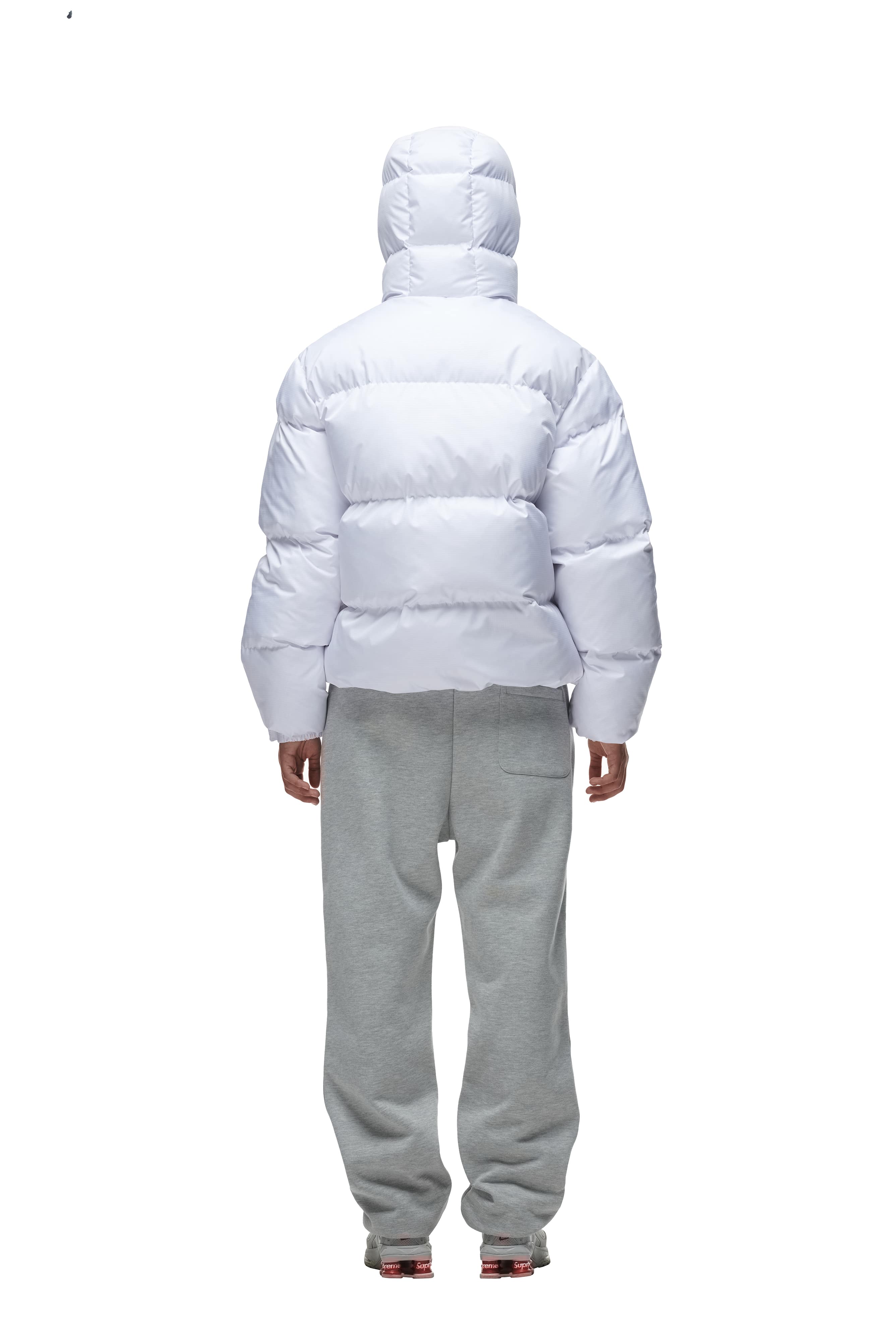 A person stands facing away against a plain white background, wearing the 6PM PUFFER JACKET WHITE with a hood, paired with gray sweatpants and sneakers.