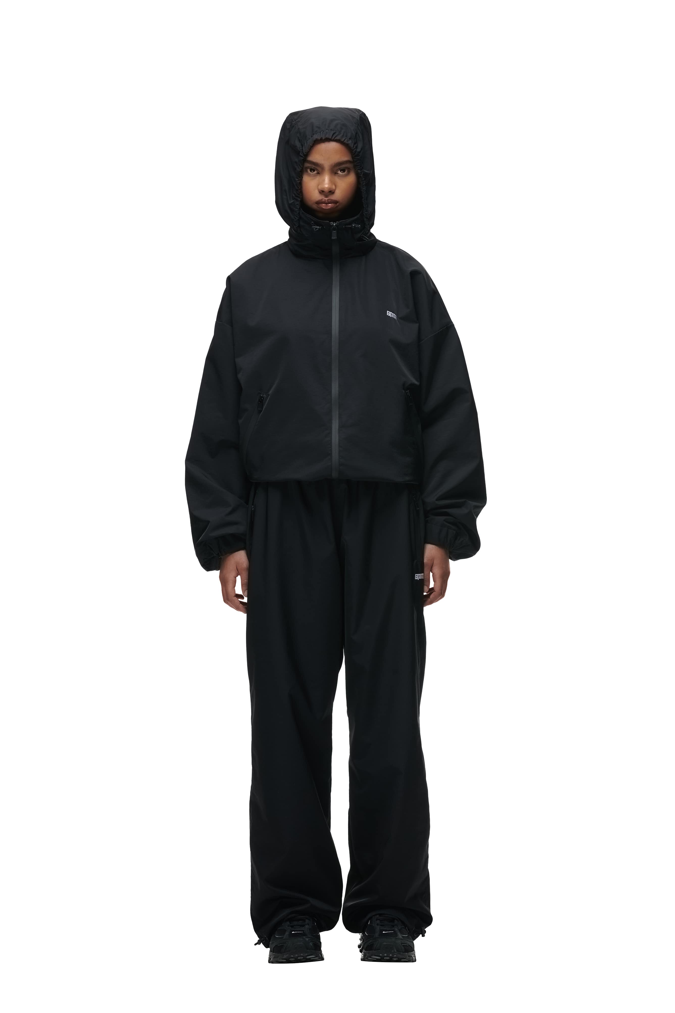 A person stands facing forward, wearing the 6PM TRACK JACKET BLACK with matching black pants. Their hands are by their sides, and they have a neutral expression. The look is sporty and casual.