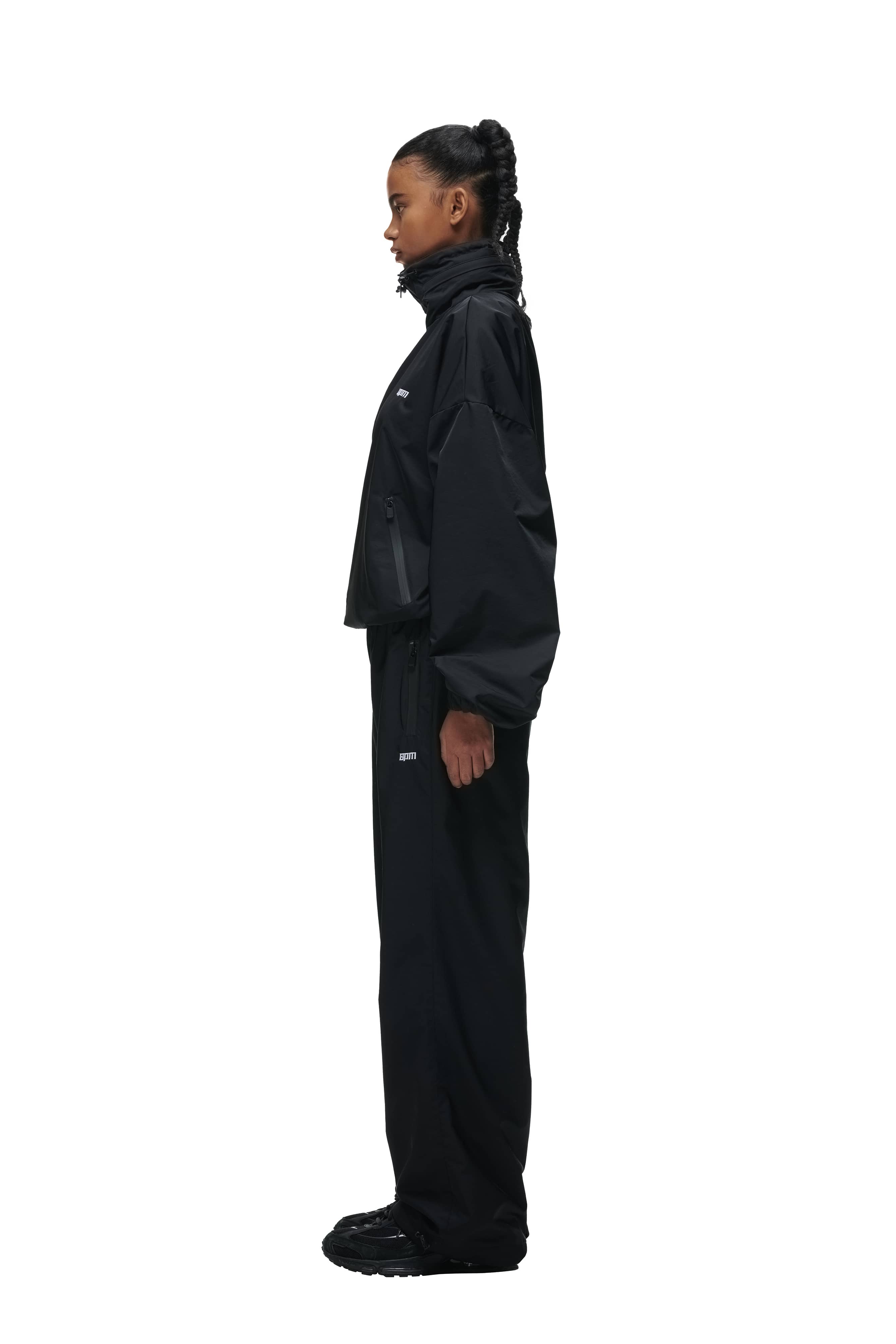 A woman stands in profile against a white background, wearing the 6PM TRACK JACKET BLACK with matching black pants and shoes. Her braided hair is tied back, and the overall look is sporty and loose-fitting.