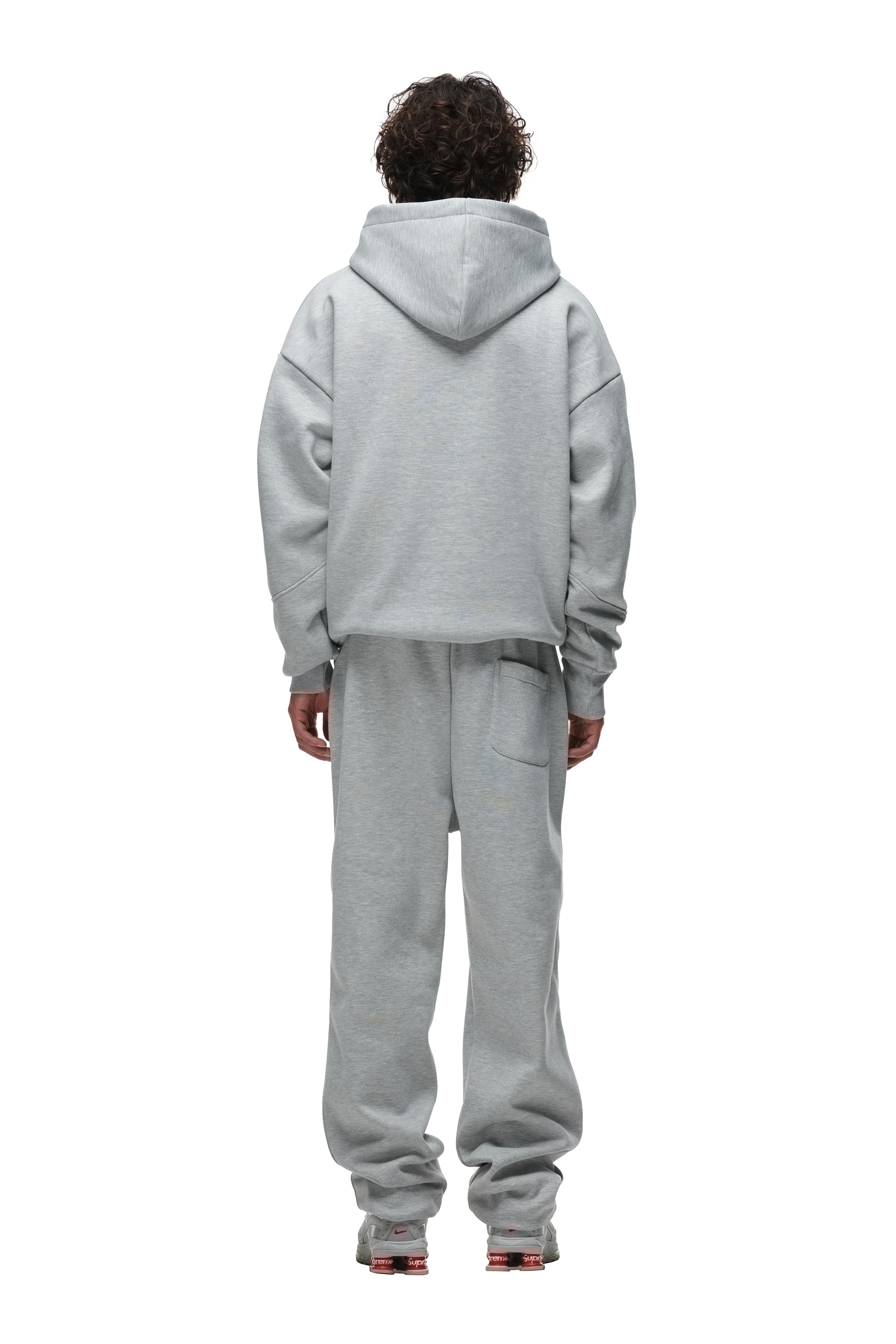 A person with curly hair faces away from the camera, wearing the 6PM DOUBLE LAYER PLAY HOODIE GREY and matching sweatpants with sneakers, set against a plain white background.