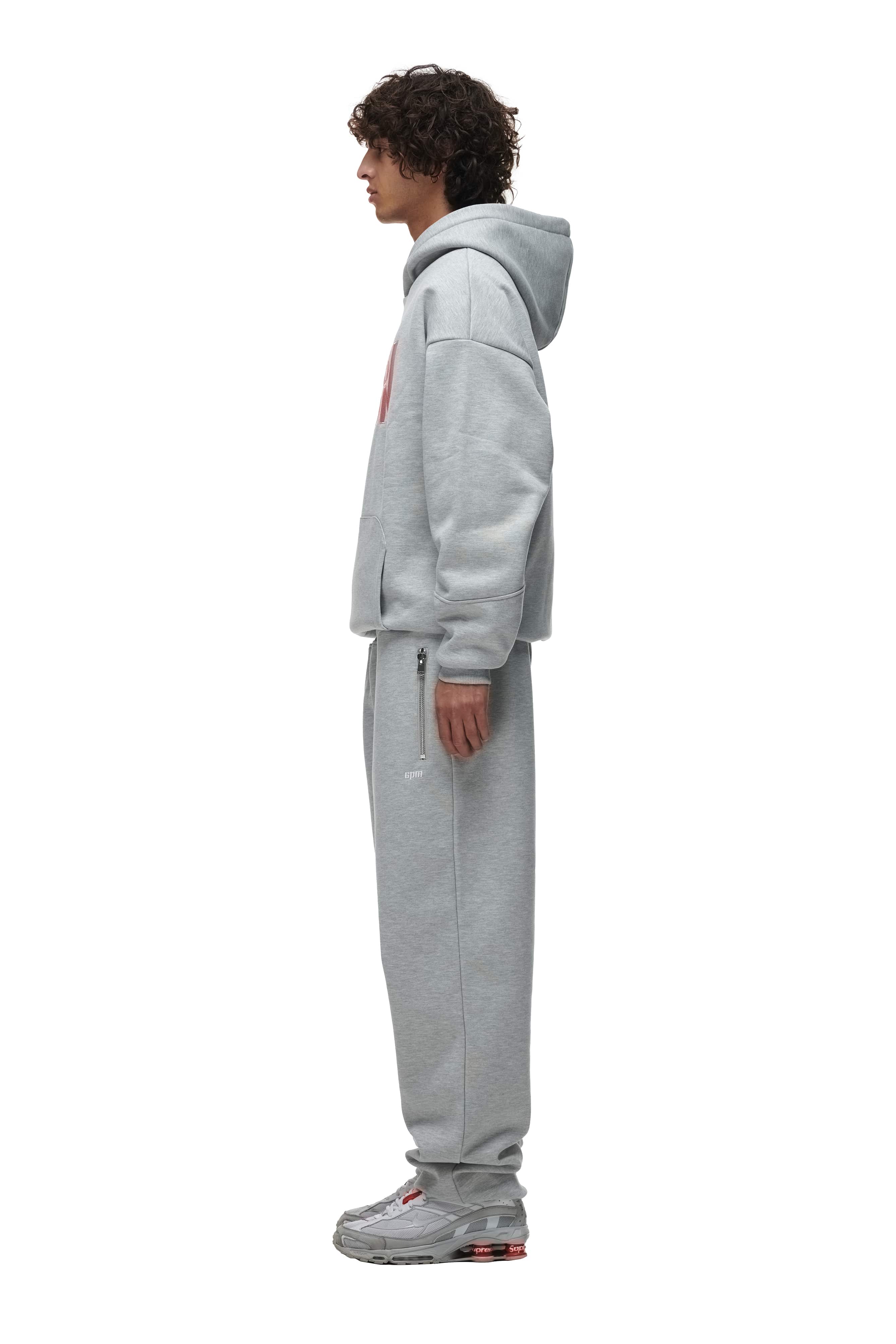 A person stands in profile wearing the 6PM DOUBLE LAYER PLAY HOODIE GREY, paired with matching sweatpants and gray sneakers, against a plain white background.