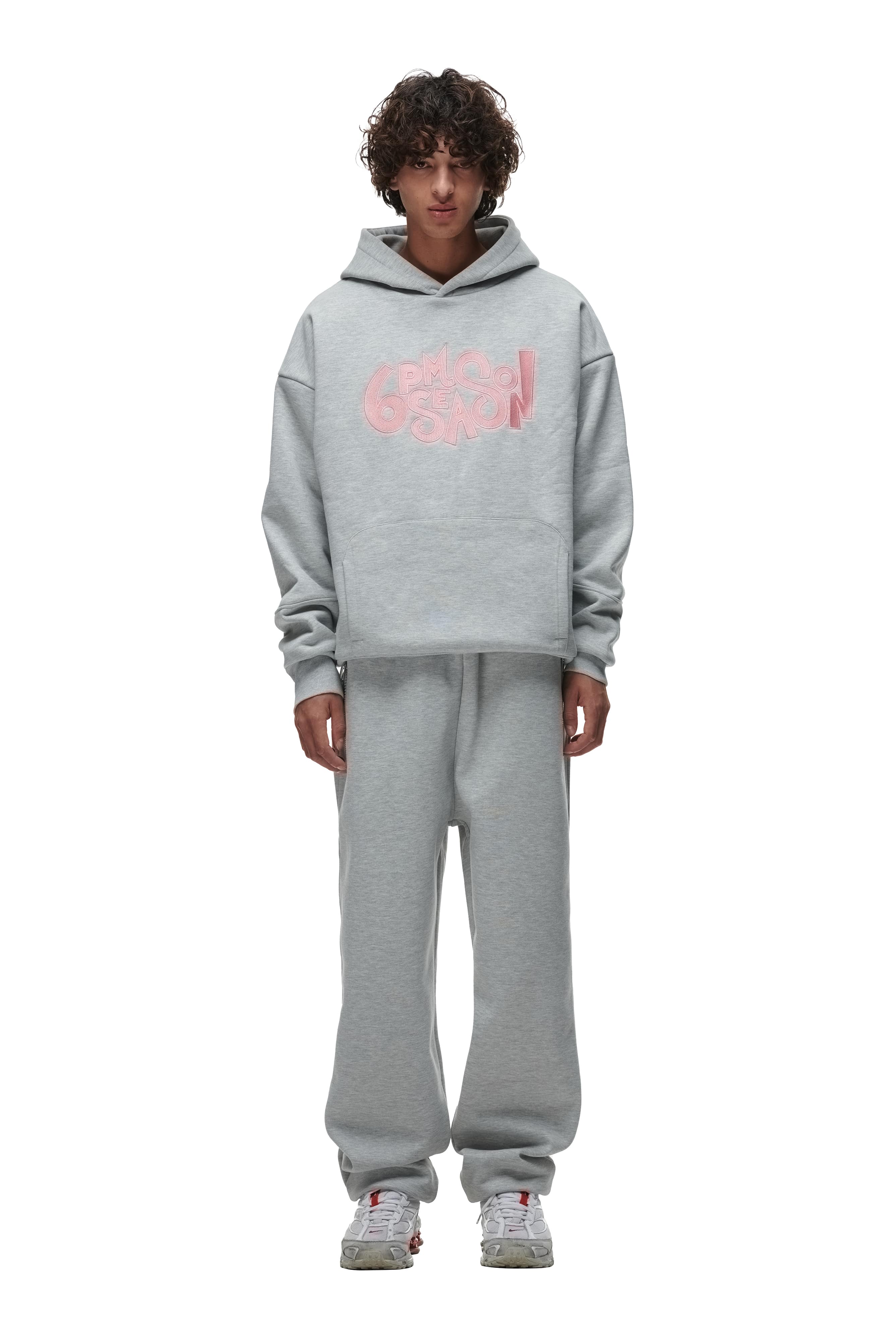 A person with curly hair wears the 6PM DOUBLE LAYER PLAY HOODIE GREY with a pink graphic on the front and matching sweatpants, standing against a white background.