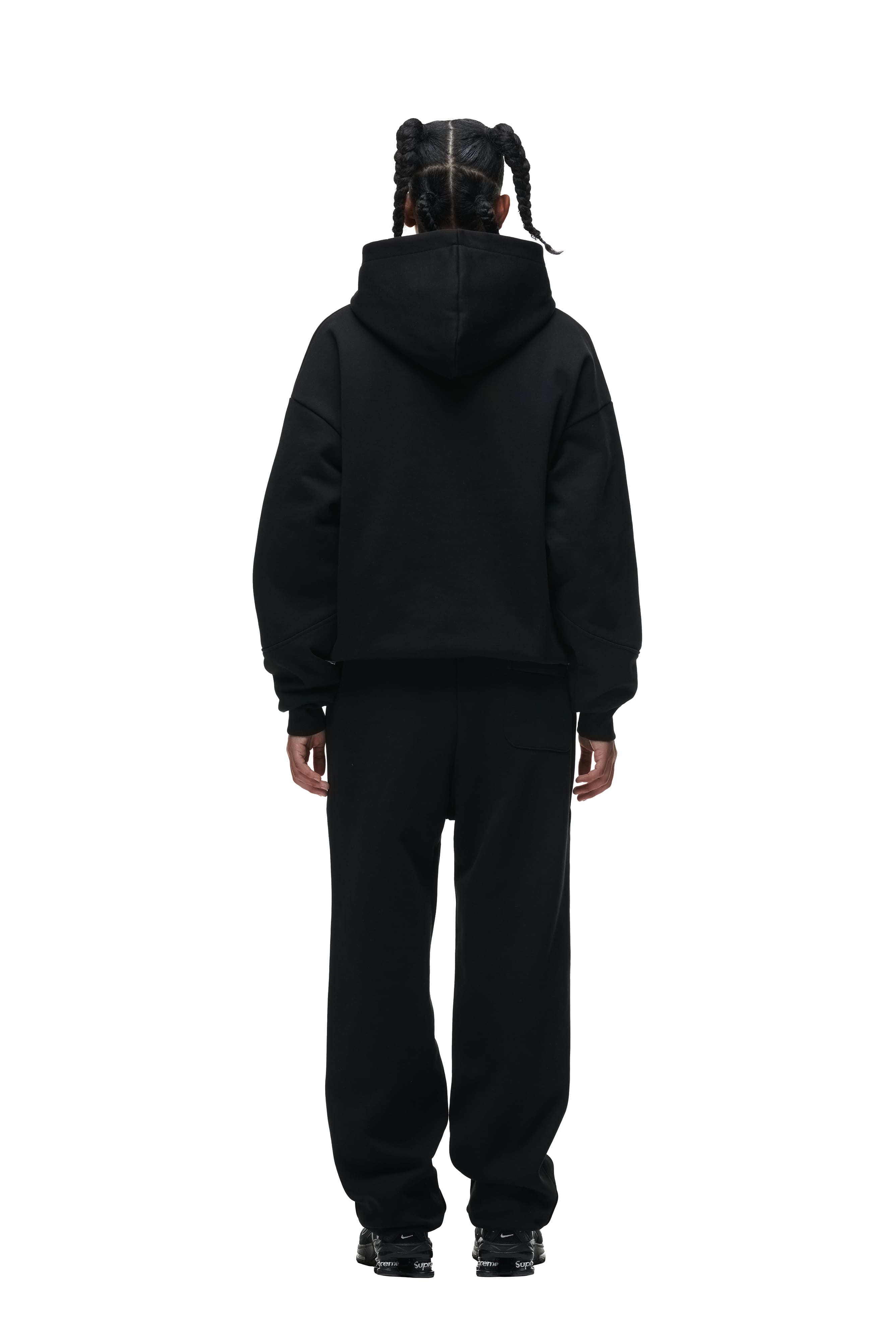 A person with braided hair faces away from the camera, wearing the 6PM DOUBLE LAYER PLAY HOODIE BLACK, black pants, and black sneakers, against a plain white background.