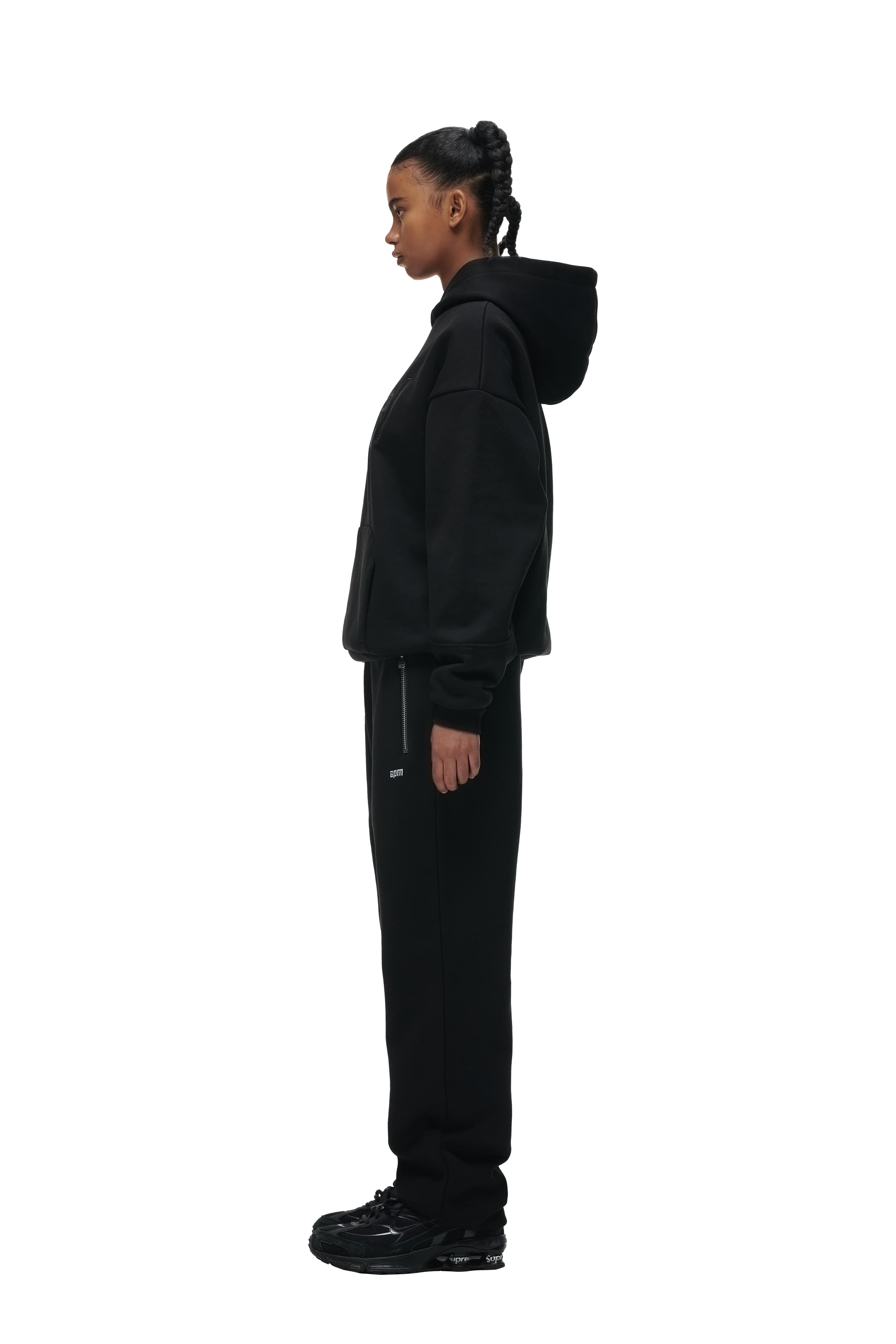 A person stands in profile facing left, wearing the 6PM DOUBLE LAYER PLAY HOODIE BLACK, along with black sweatpants and black sneakers, against a plain white background.