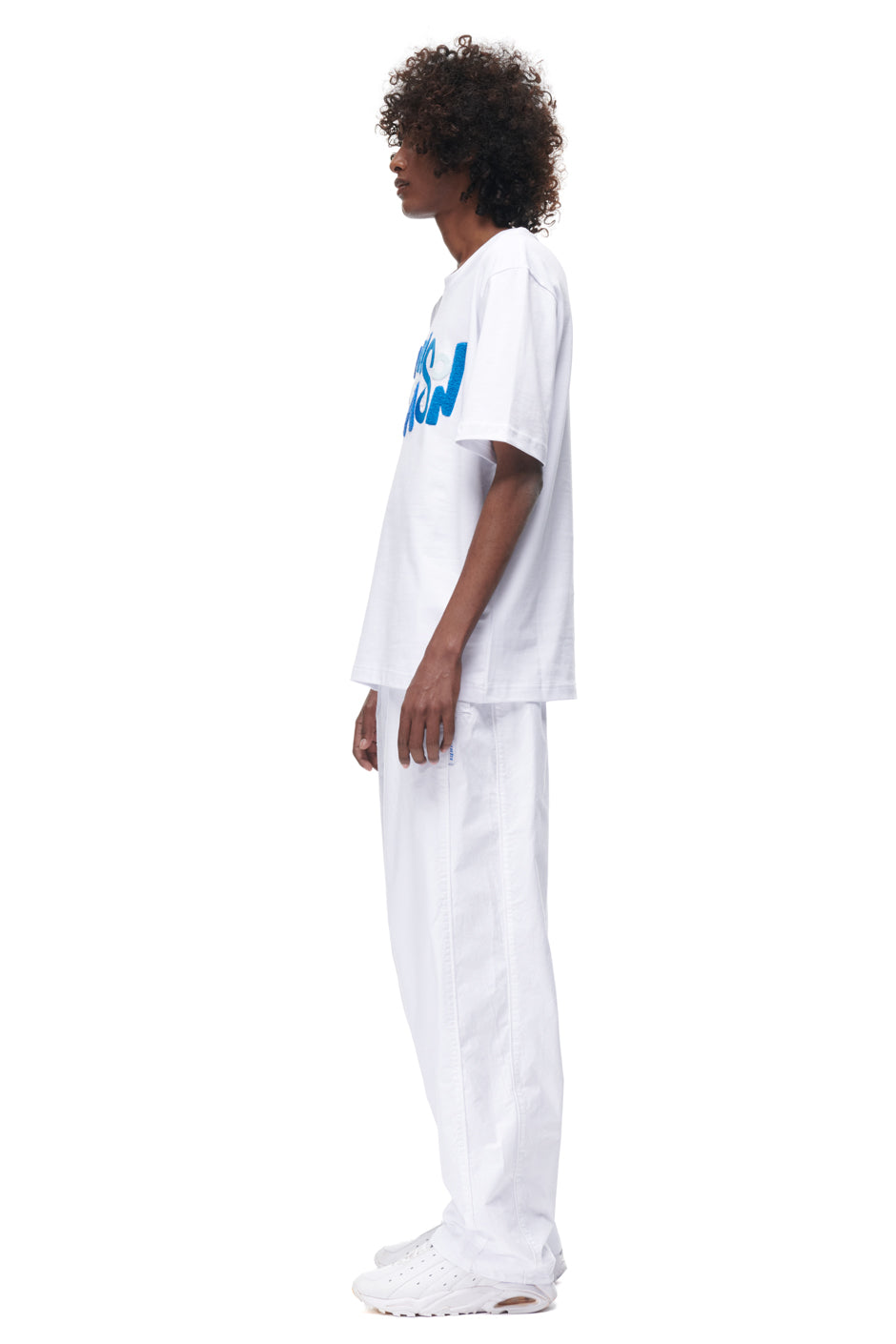 A person with curly hair stands in profile against a plain white background, wearing the 6PM PLAY T-SHIRT WHITE, paired with white pants and white sneakers.