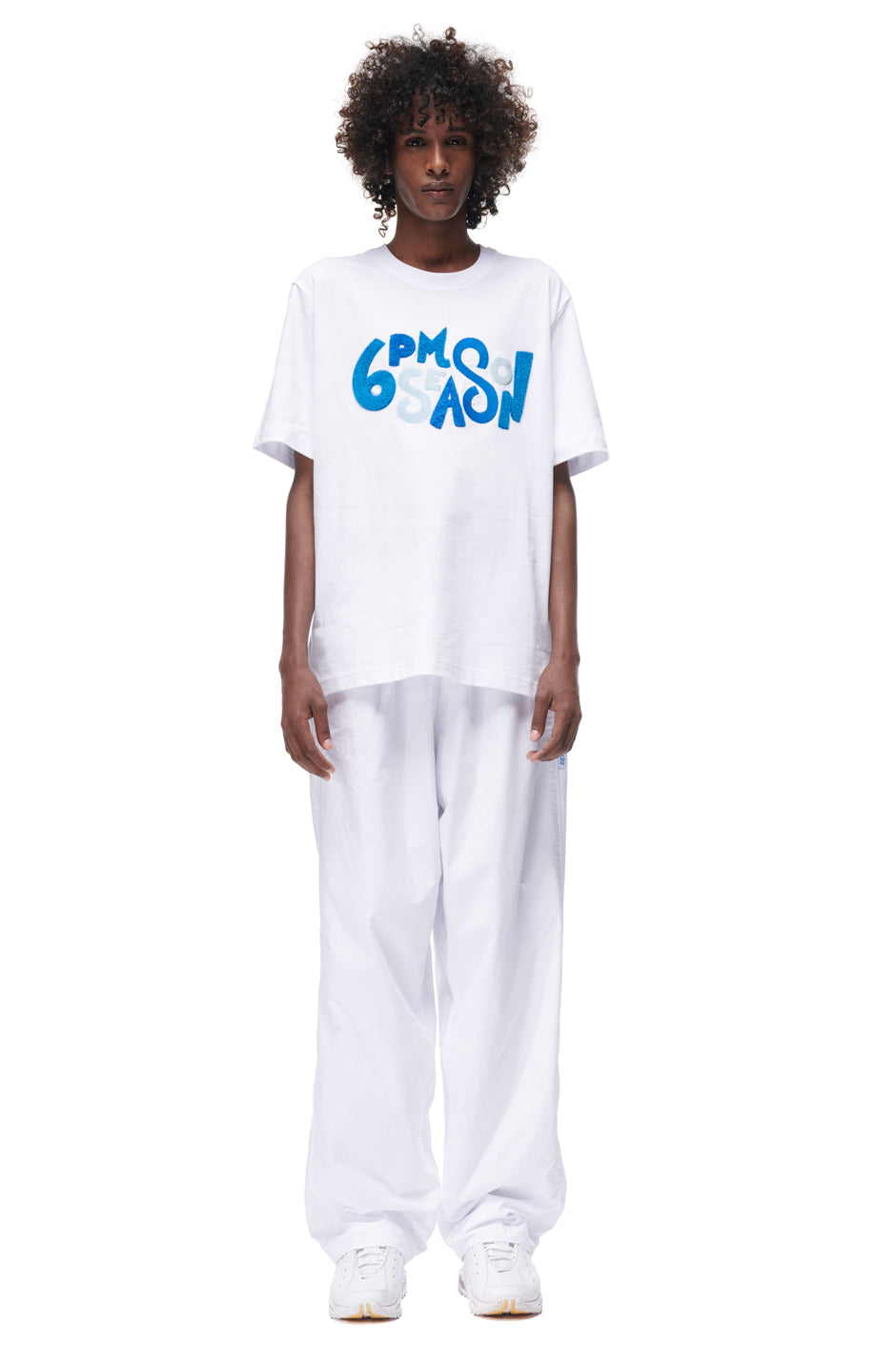 A person with curly hair wears the 6PM PLAY T-SHIRT WHITE, featuring bold blue "6PM SEASON" text, and loose white pants, posing against a plain white background.