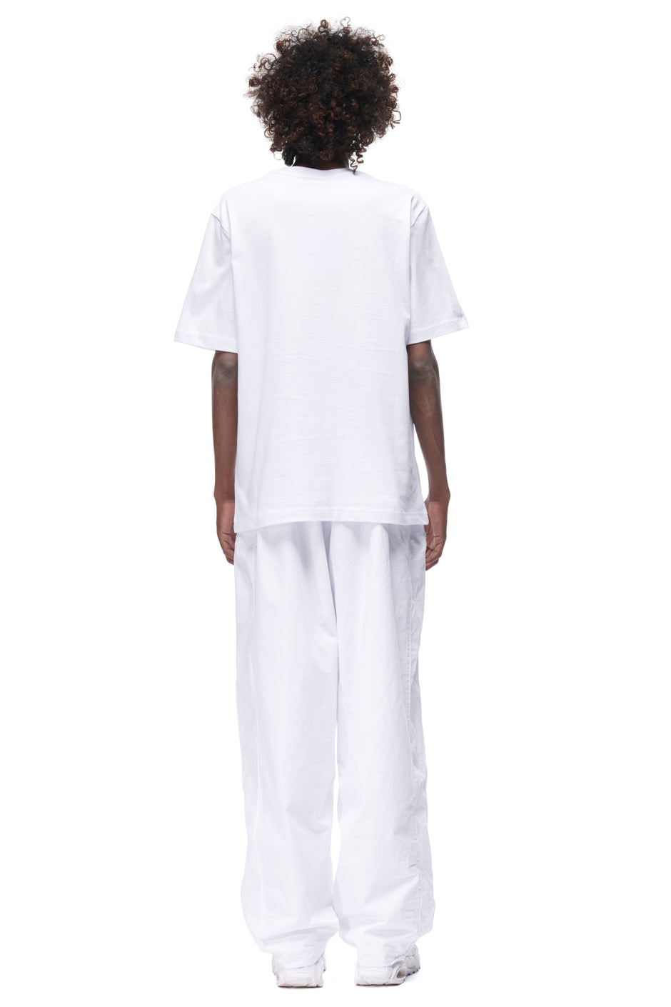 A person with curly hair stands facing away against a plain white background, wearing the 6PM PLAY T-SHIRT WHITE, loose white pants, and white shoes.