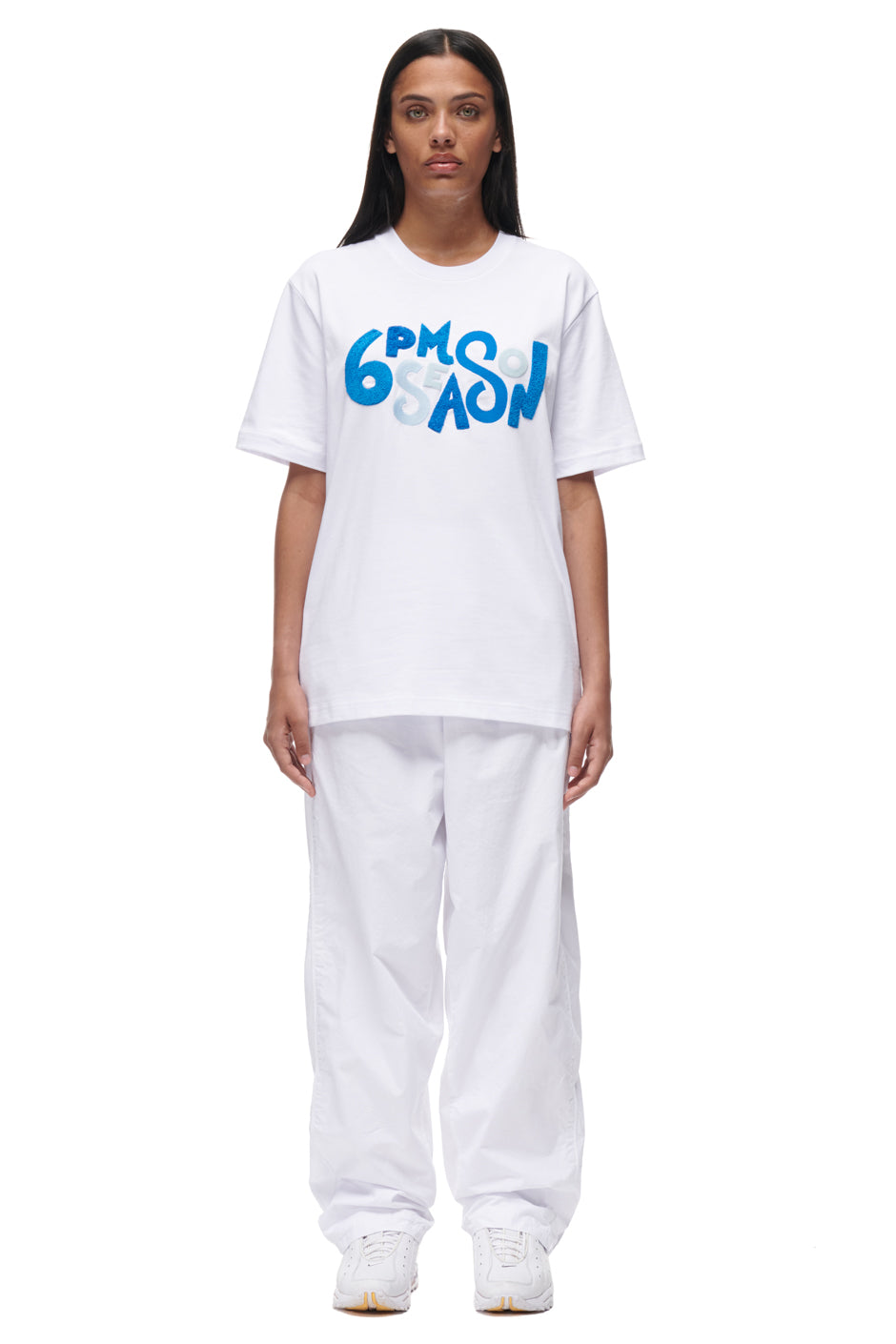 A woman faces forward against a plain white background, wearing the 6PM PLAY T-SHIRT WHITE with bold blue lettering, paired with baggy white pants and white sneakers.