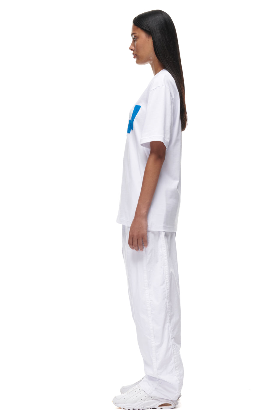 A woman in profile stands against a white background, wearing the 6PM PLAY T-SHIRT WHITE, paired with white baggy pants and sneakers. Her straight dark hair is worn down.