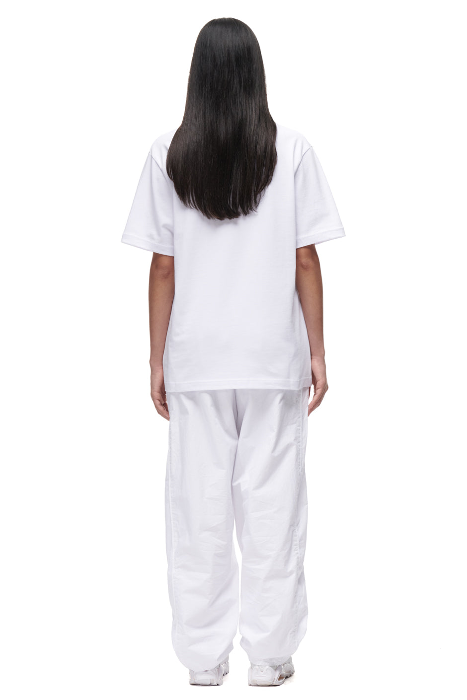 A person with long, straight black hair stands facing away, wearing the 6PM PLAY T-SHIRT WHITE, baggy white pants, and white shoes against a plain white background.