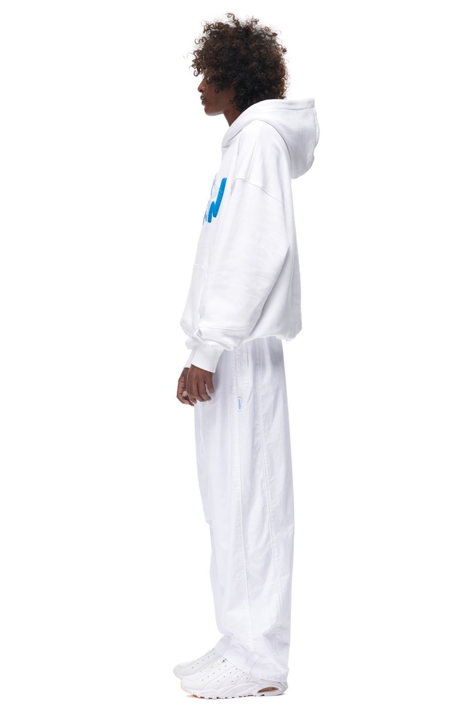 A person stands in profile wearing the 6PM PLAY HOODIE WHITE, paired with white baggy pants and white sneakers, set against a plain white background.
