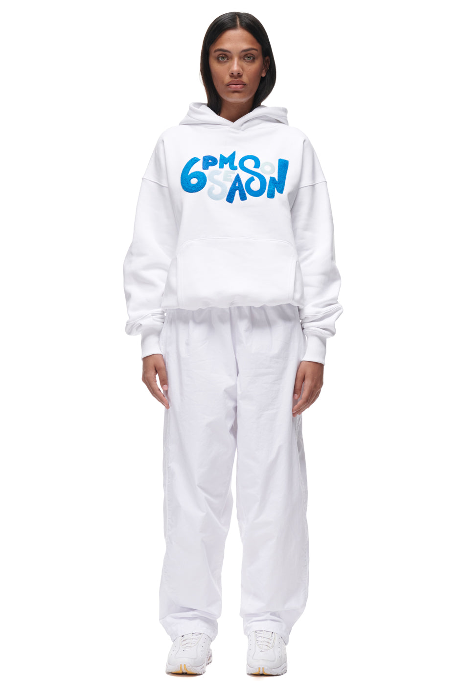 A woman with straight dark hair and a neutral expression wears the 6PM PLAY HOODIE WHITE, an all-white hoodie with 6PM SEASON in bold blue letters on the front, facing forward.